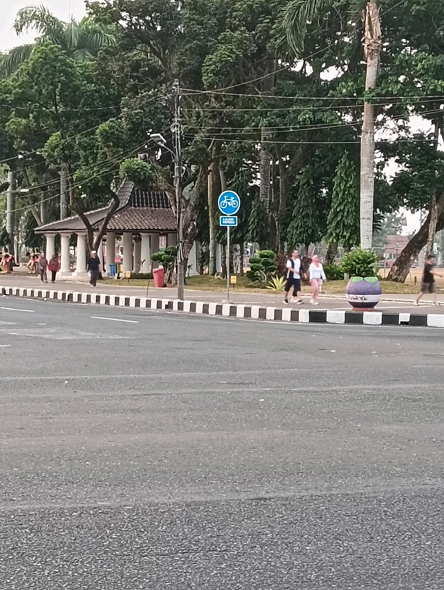The image depicts a street scene in Purworejo, Indonesia, likely in a park or recreational area given the presence of trees, a gazebo-like structure, and walking paths. The foreground shows a paved road with lane markings and a distinct black and white striped curb separating it from a pedestrian walkway. On the walkway, several individuals are visible. Two women, one in a white top and pink pants, the other in a black and white top and dark shorts, are walking together. Another man in a black shirt and shorts is seen walking away from the camera. Further back, near a pavilion with white pillars and a tiled roof, a man in a dark shirt and cap is walking. Several other figures, likely pedestrians, are also present along the path. A blue sign with a bicycle icon and text below it is mounted on a pole near the walkway. A large, dark purple planter shaped like a fruit, possibly a mangosteen, sits on the edge of the walkway. Red and blue trash receptacles are also visible. Lush green trees, including palm trees, form a dense canopy behind the walkway, creating a shaded environment. Power lines and a street light are also present in the background. The overall impression is a public space being utilized by pedestrians on a walk.