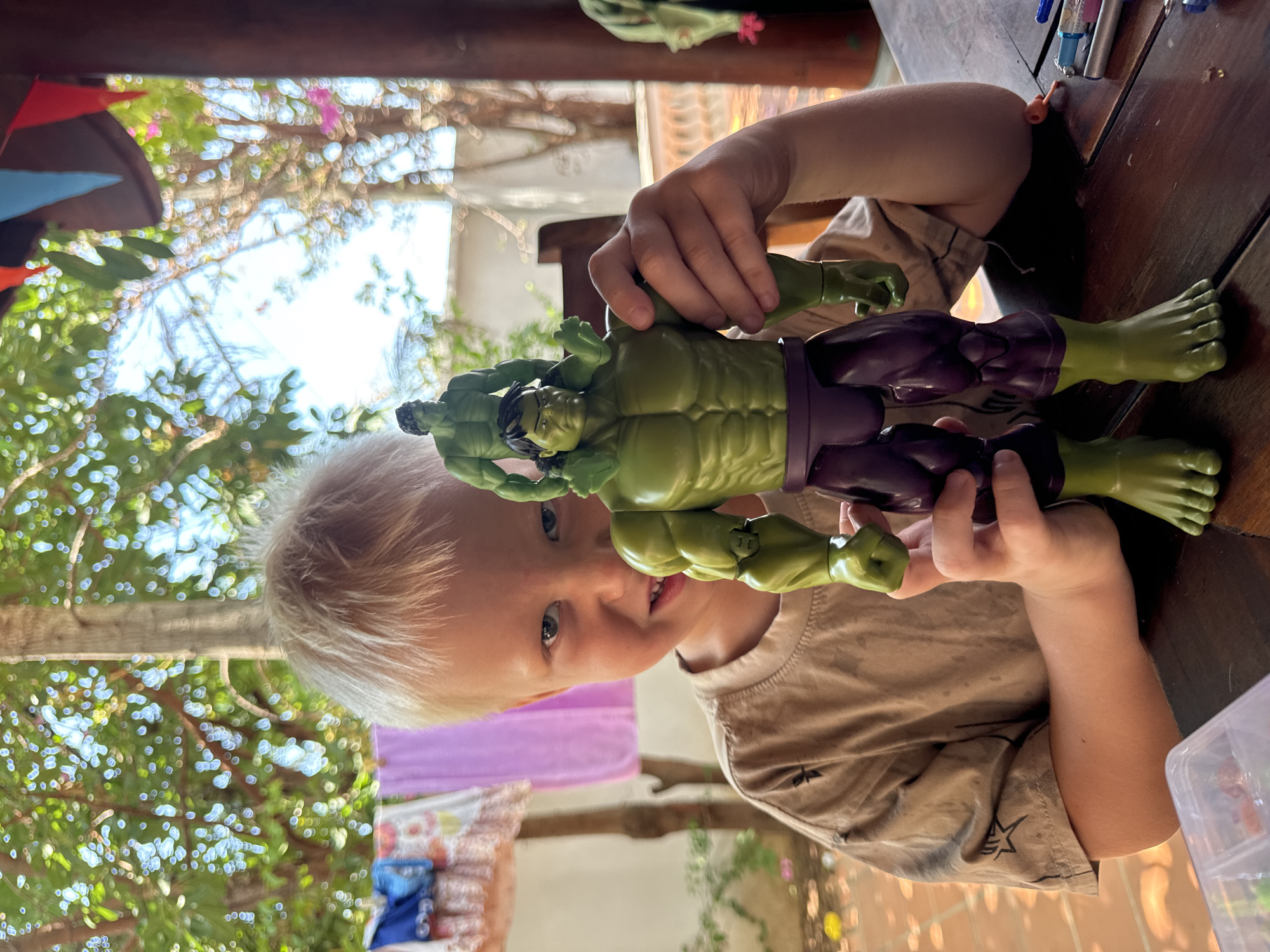 A young, fair-skinned child with blond hair holds up two Hulk action figures. The child is wearing a beige shirt with black stars. Behind the child, laundry is hanging, including a purple towel and items with floral patterns and blue accents. Sunlight filters through trees in the background. The child is indoors, sitting at a wooden table where pens and other items are located. While the general scene is established, there is insufficient information in the image to specify the exact location within Mũi Né, Vietnam.