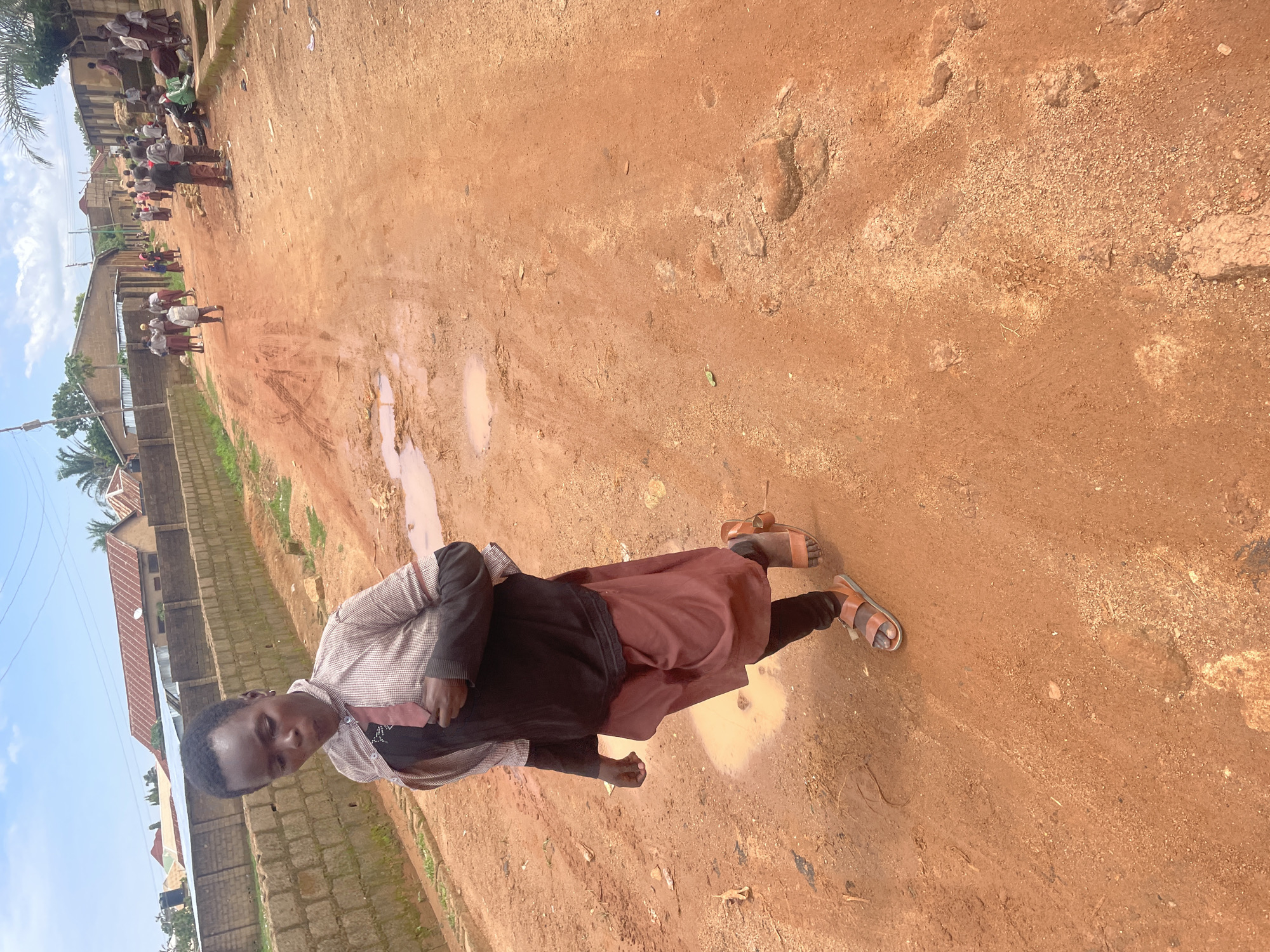 In Barakin Akawo, Nigeria, a young person walks down a reddish-brown dirt road. They are wearing a black t-shirt under a light-colored, patterned button-down shirt with a dark tie. A brown skirt or wrap covers their legs, and they are wearing dark leggings and brown sandals. The road is uneven with several puddles of water and tire tracks. In the background, several people are visible, some walking and others standing or sitting. Buildings with corrugated metal roofs and brick walls line one side of the road, and vegetation can be seen in the distance. The sky is partly cloudy.
