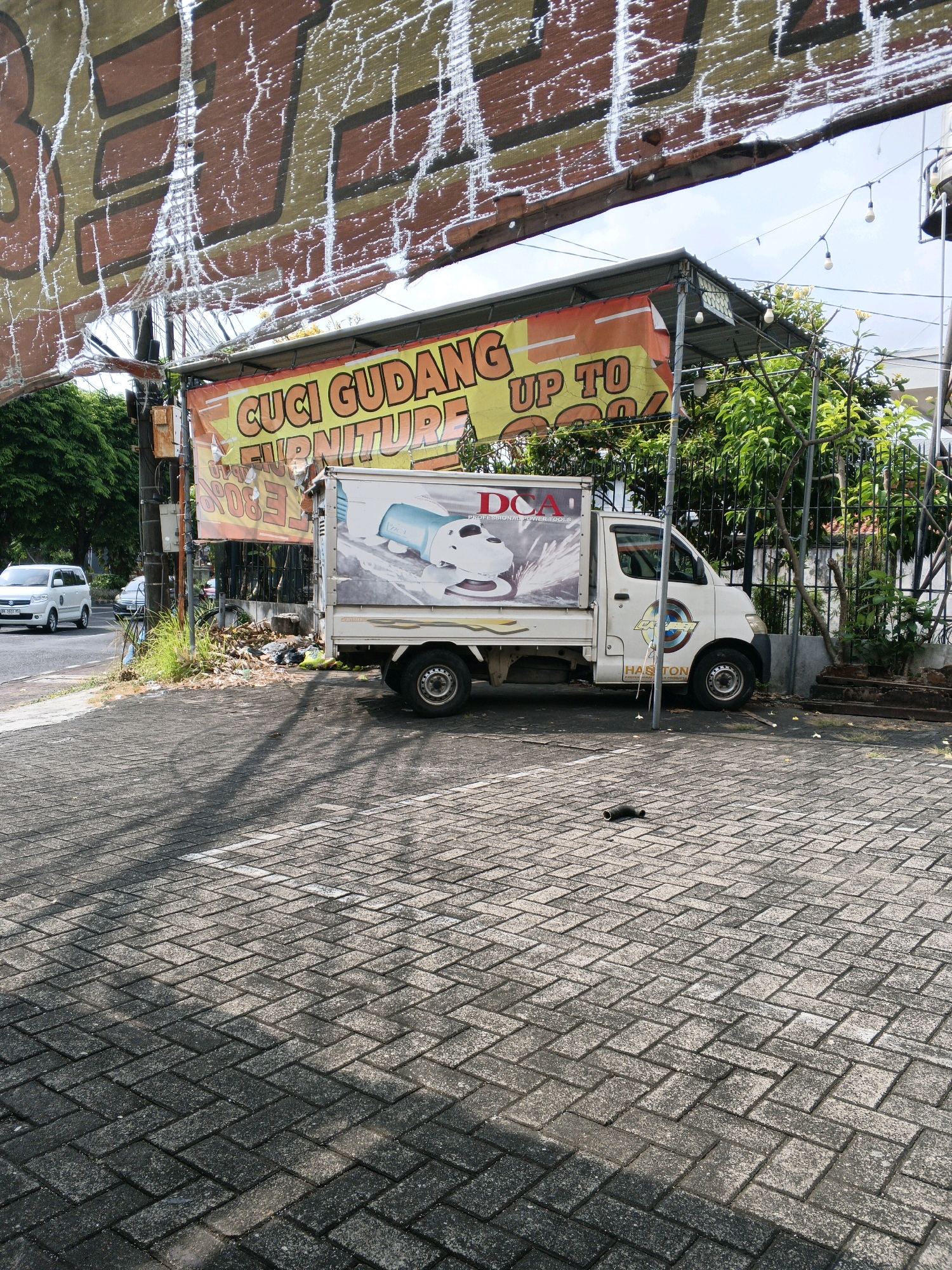 The image shows a white pickup truck parked on a cobblestone street in Denpasar, Indonesia. The truck has a large advertisement on its side for "CUCI GUDANG FURNITURE UP TO 50%". The truck is parked under a canopy with a banner that also advertises the furniture sale. In the background, there are trees and a fence. The sky is visible, and there are some street lights hanging from wires. The scene appears to be on a street in a commercial area.