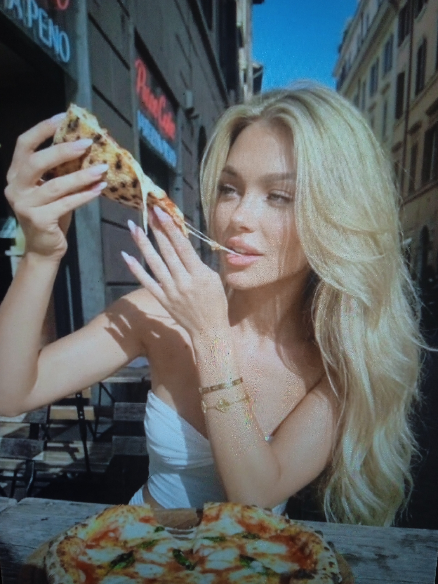 An unidentifiable young woman with long blonde hair is holding a slice of pizza, which has a long string of cheese stretching from it to her mouth. She is wearing a white strapless top and a gold bracelet. In the foreground, a whole pizza is visible on a wooden table. The background appears to be a street scene with buildings and a blue sky. The image has a grainy quality. There is no discernible information in the image to indicate the location as Chernivtsi, Ukraine.