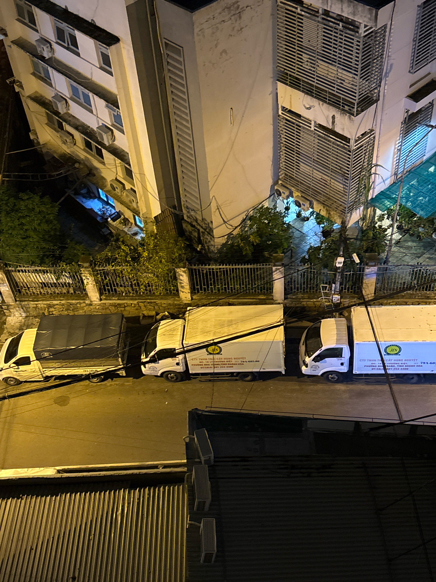In Nha Trang, Vietnam, at night, three delivery trucks are parked on a street adjacent to a multi-story building. Two white trucks with covered cargo areas are positioned next to each other, with a third white truck on the far left also with a covered cargo area. The building has multiple windows, some with air conditioning units attached. A metal fence with vertical bars runs along the base of the building, separating it from the street. Overgrown vegetation is visible behind the fence. Electrical wires are strung across the scene. The street is paved, and the lighting suggests an urban environment.