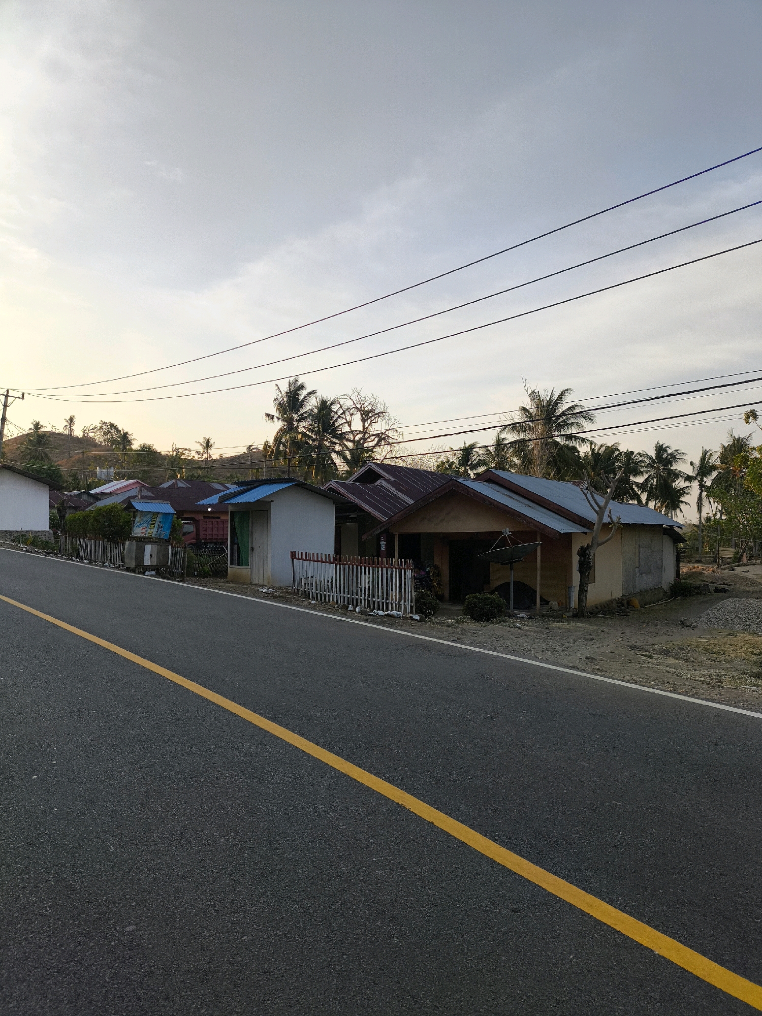 The image depicts a rural roadside scene in Indonesia. A paved road with a yellow center line runs diagonally from the bottom left towards the center right of the frame. Along the right side of the road, several modest houses with corrugated metal roofs are visible. These houses appear to be single-story and are situated on a slightly elevated, unpaved area. Palm trees and other tropical vegetation are scattered behind and around the houses, suggesting a warm climate. The sky is overcast with a pale, hazy quality, indicating either early morning or late afternoon light. Overhead, several dark power lines crisscross the sky, supported by utility poles visible on the left side of the image. The overall impression is one of a simple, functional settlement in a tropical environment. There are no people or animals present in the scene.