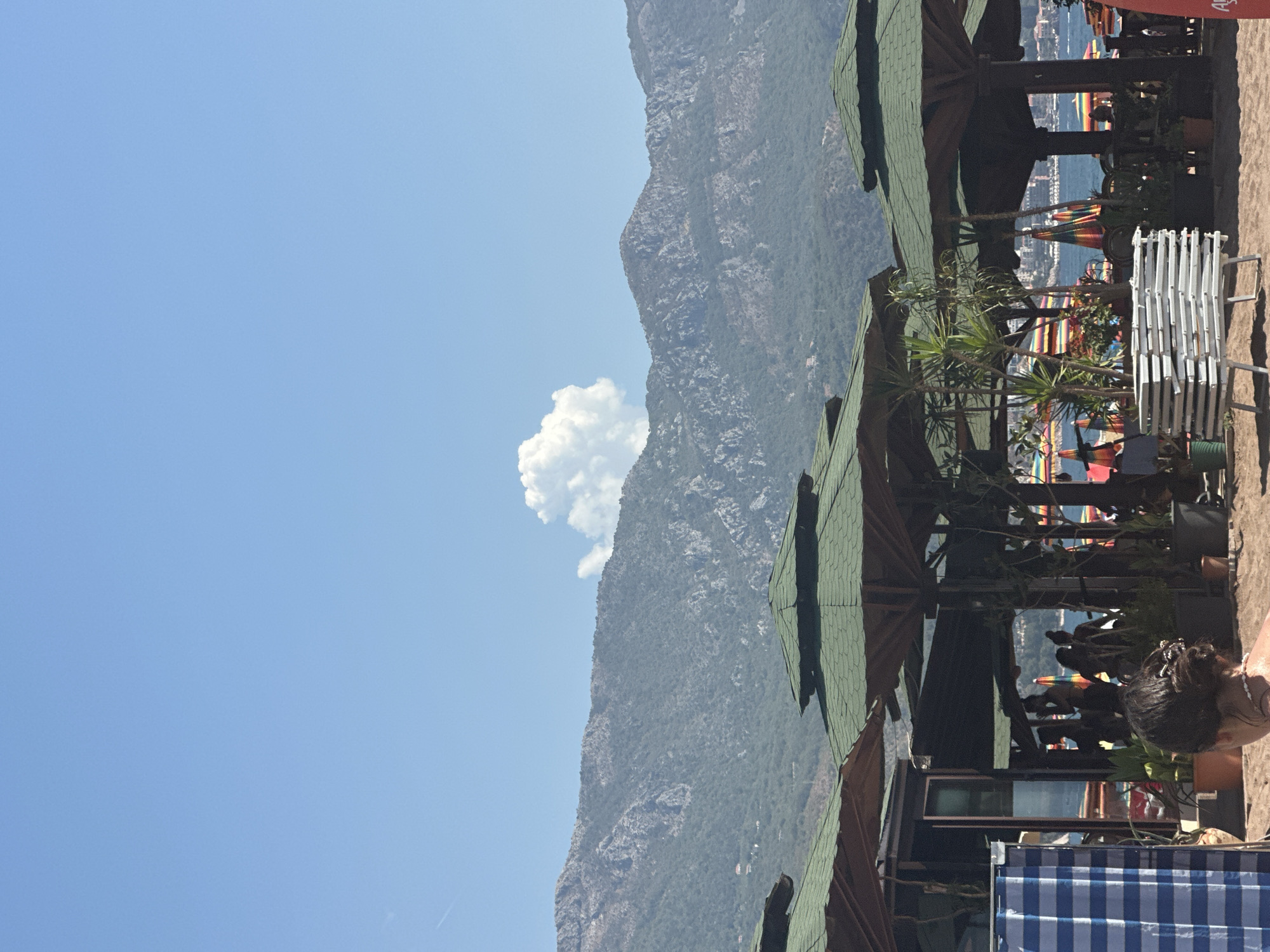 The image depicts a sunny day at a beach in Boreti, Montenegro. In the foreground, a woman with her back to the camera, her hair tied up with accessories, is partially visible. To her left, a blue and white striped privacy screen stands. Behind this, a row of thatched-roof umbrellas provides shade over what appears to be a beachside establishment. Plants in pots are scattered around the seating areas under the umbrellas. In the middle ground, several people are visible in silhouette through the open structure of the umbrellas, some appearing to be patrons or staff. Beyond them, the turquoise sea is visible, dotted with colorful beach umbrellas. Further in the distance, a city skyline is faintly discernible along the coast. The background is dominated by a range of large, green, and rocky mountains under a clear blue sky with a single, fluffy white cloud. The overall scene conveys a relaxed atmosphere of a summer day at the beach.
