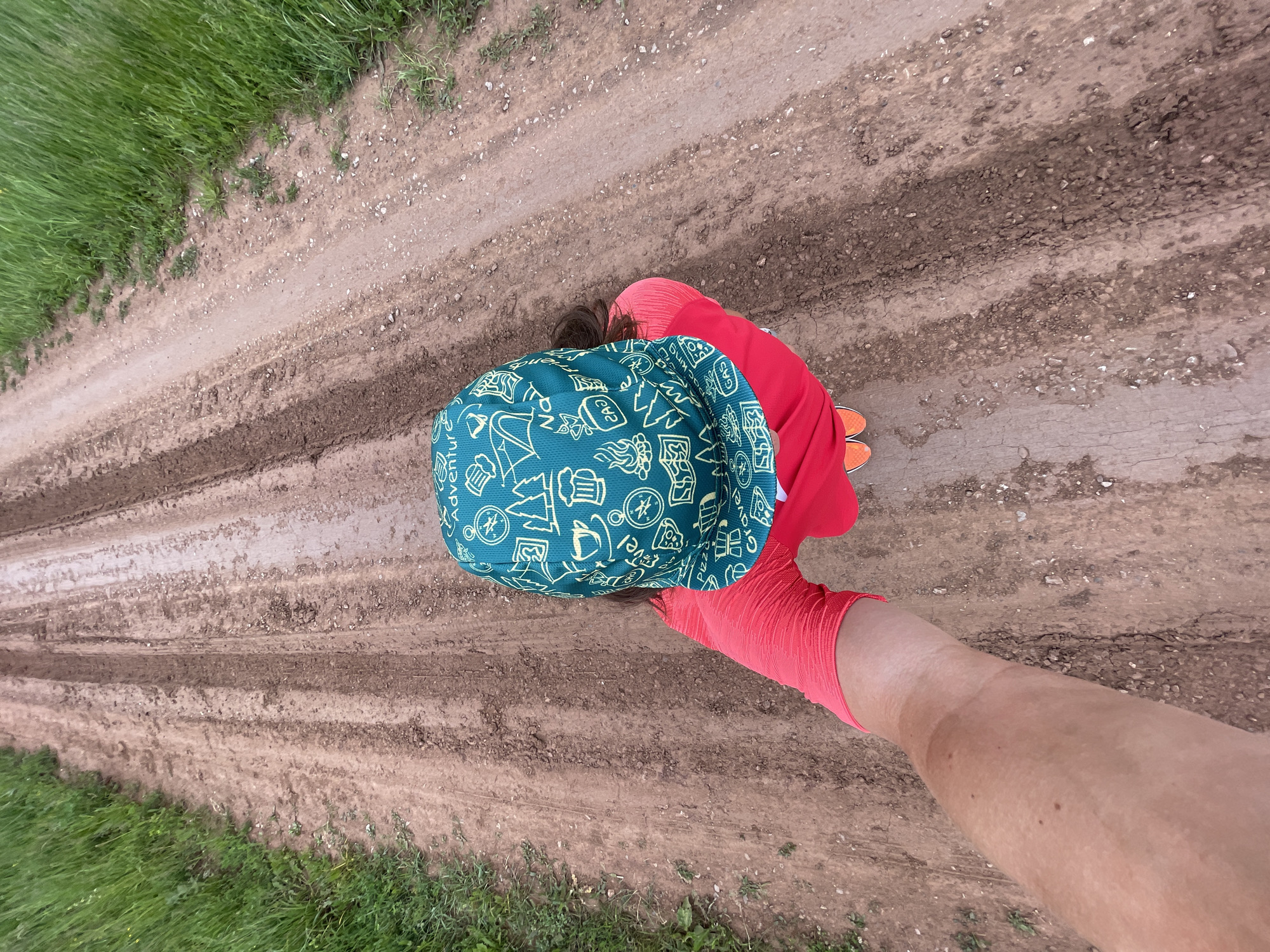 An overhead, point-of-view shot shows a person from the shoulders up from the chest down. The person is wearing a red long-sleeved shirt and an orange baseball cap with a teal brim and patterned crown. The person is standing on a dirt road that has tire tracks and grass on either side. The road goes into the distance.