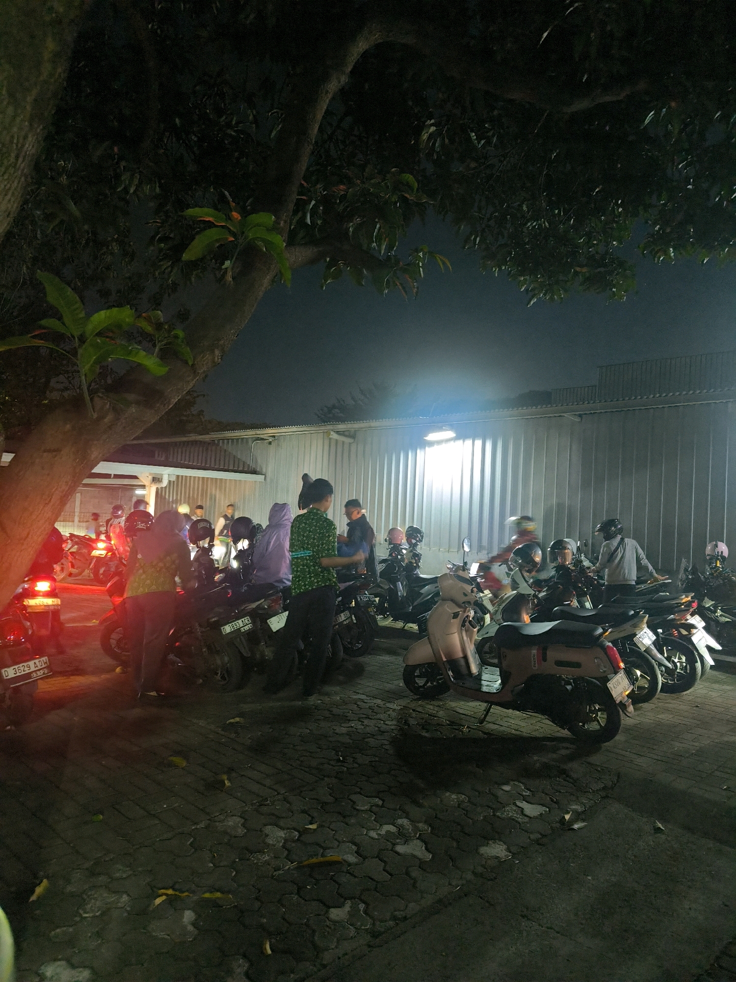 The image depicts a nighttime scene of a parking area filled with motorcycles, possibly located in an outdoor commercial or residential area in Bandung City, Indonesia. Several individuals are visible, some standing and conversing, while others appear to be preparing to depart on their motorcycles. The environment includes a large tree with branches extending into the frame, a building with a corrugated metal facade, and a paved ground surface. The lighting suggests artificial illumination, likely from streetlights or building lights, casting shadows and highlighting the vehicles and people. The overall atmosphere is one of activity and gathering, common in urban Indonesian settings during the evening.