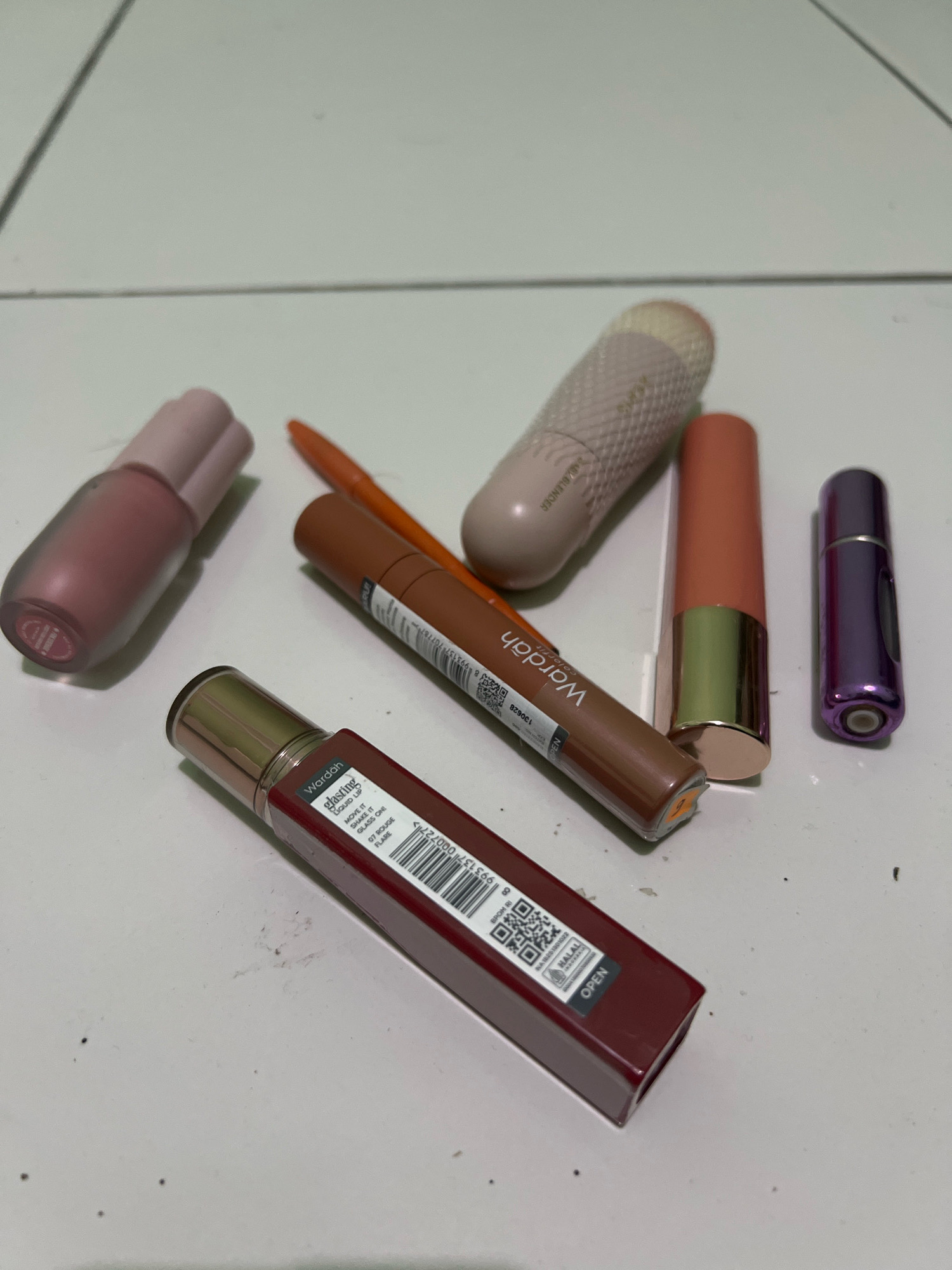 The image displays an overhead view of a collection of makeup products on a light-colored, tiled surface, possibly a floor. The items are arranged loosely, with some overlapping.
From left to right, the objects include:
1. A matte pink lipstick tube with a slightly lighter pink cap. The cap appears to be twisted off and placed behind the tube.
2. An orange eyeliner pencil.
3. A light beige, textured cylindrical object, possibly a makeup blender or a similar tool, with a lighter beige tip. The word "BLENDER" is faintly visible on its side.
4. A matte coral-pink lipstick tube with a gold band at the bottom.
5. A brown liquid lipstick tube with a gold cap and applicator. The label indicates "Wardah" and "glasting liquid lip" with a shade name "07 ROUGE FLAME".
6. A small, metallic purple cylindrical object, likely a travel-sized perfume atomizer or a similar cosmetic container.
7. A dark red liquid lipstick tube with a gold cap. The label shows "Wardah" and "glasting liquid lip" and a barcode. It also has a sticker that says "OPEN".
The environment appears to be indoors, with a white tiled floor that has visible grout lines. There are small dark specks scattered on the surface, suggesting dust or debris. The lighting is diffused, creating soft shadows. The image does not provide any specific contextual clues to pinpoint its exact location in Tlogoadi, Indonesia, beyond the nature of the objects themselves being common cosmetic items.