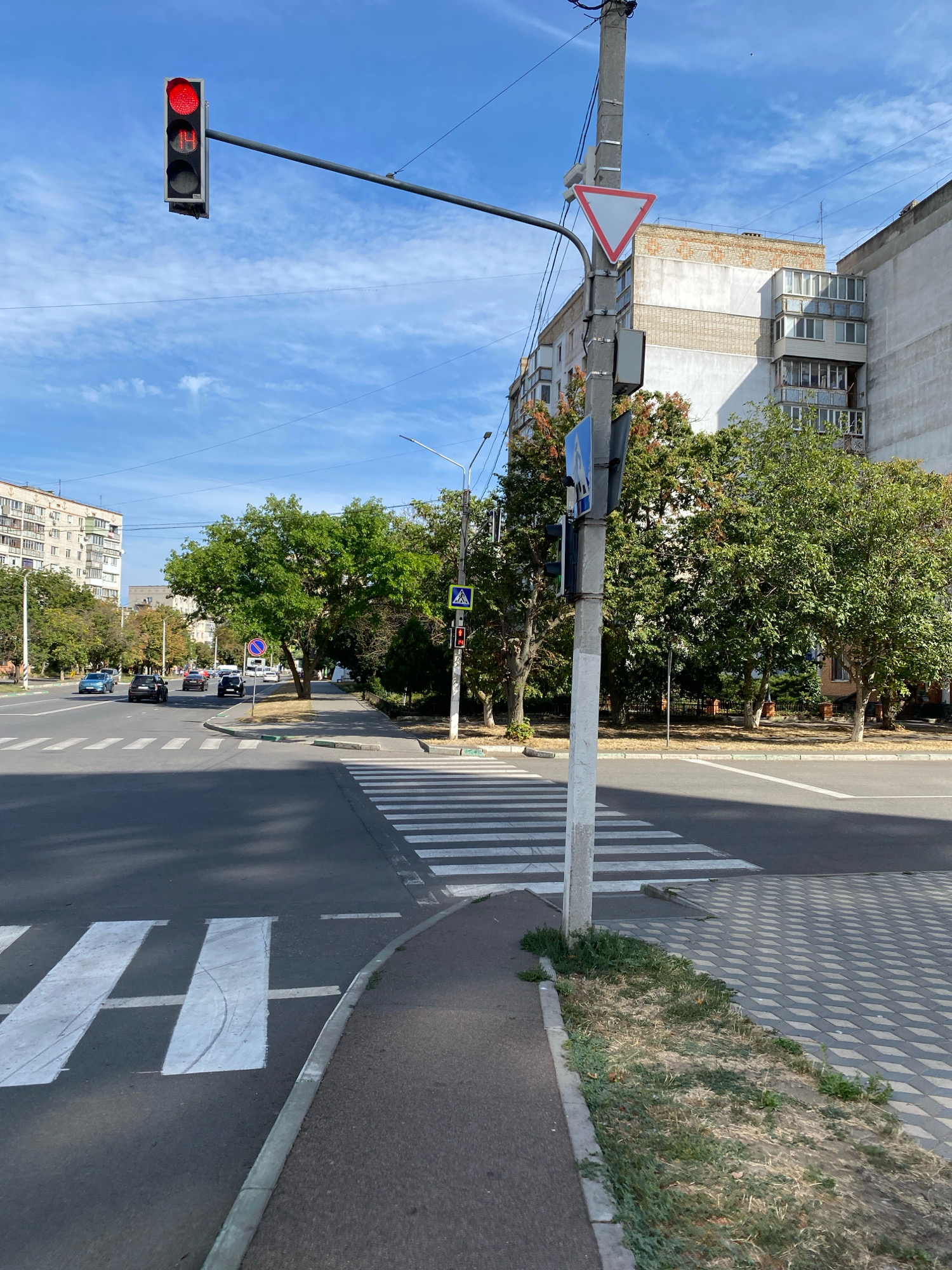 The image depicts a street scene in Chornomorsk, Ukraine, under a clear blue sky with scattered clouds. A traffic light displays a red signal, indicating a stop. A pedestrian crossing is clearly marked with white stripes on the asphalt. To the right of the crossing, a sidewalk runs alongside the road, bordered by a patch of dry grass and vegetation. Beyond the sidewalk, a street lamp stands tall, accompanied by traffic signs. In the background, apartment buildings rise, characteristic of urban architecture. Trees with lush green foliage line the street, adding to the cityscape. Several cars are visible on the road, navigating the intersection. The overall impression is of a typical urban environment in Ukraine on a bright day.