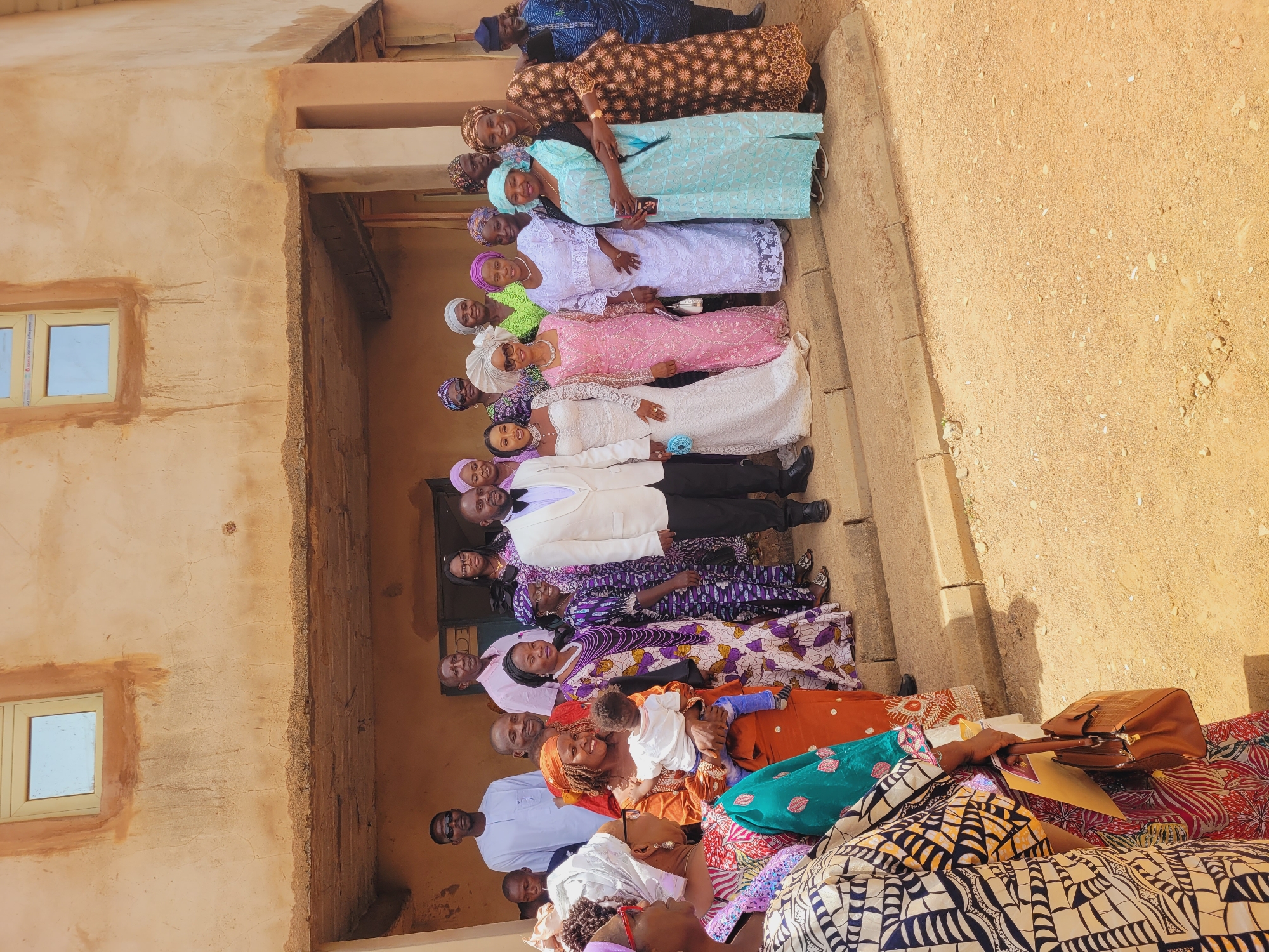 An outdoor scene in Bukuru, Nigeria, captures a large group of approximately 30-35 individuals posing for a photograph in front of a light brown, two-story building. The building features two rectangular windows with light-colored frames on the upper level and a shaded ground-floor entrance area supported by pillars. The ground in the foreground is unpaved, sandy-colored earth with scattered small stones, leading up to concrete steps where the group is primarily positioned.
At the center are a man and a woman, presumably a groom and bride. The man wears a white suit jacket, a black bow tie, and black trousers. The woman wears a long, white, intricate lace wedding gown and holds a small light blue object. Both are smiling.
The surrounding individuals are mostly adults, consisting of a mix of men and women. The women are dressed in a variety of colorful, often patterned, traditional Nigerian attire including long dresses and matching headwraps (gele), in shades of purple, pink, brown, light blue, and various ankara prints. Several women are smiling. One woman on the left holds an infant dressed in a white top and blue pants. Another woman wears a large white hat and sunglasses. The men wear traditional robes (agbada) in colors such as white, light pink, and dark blue, or casual shirts. All individuals face towards the camera, arranged in multiple rows on the steps and ground level.