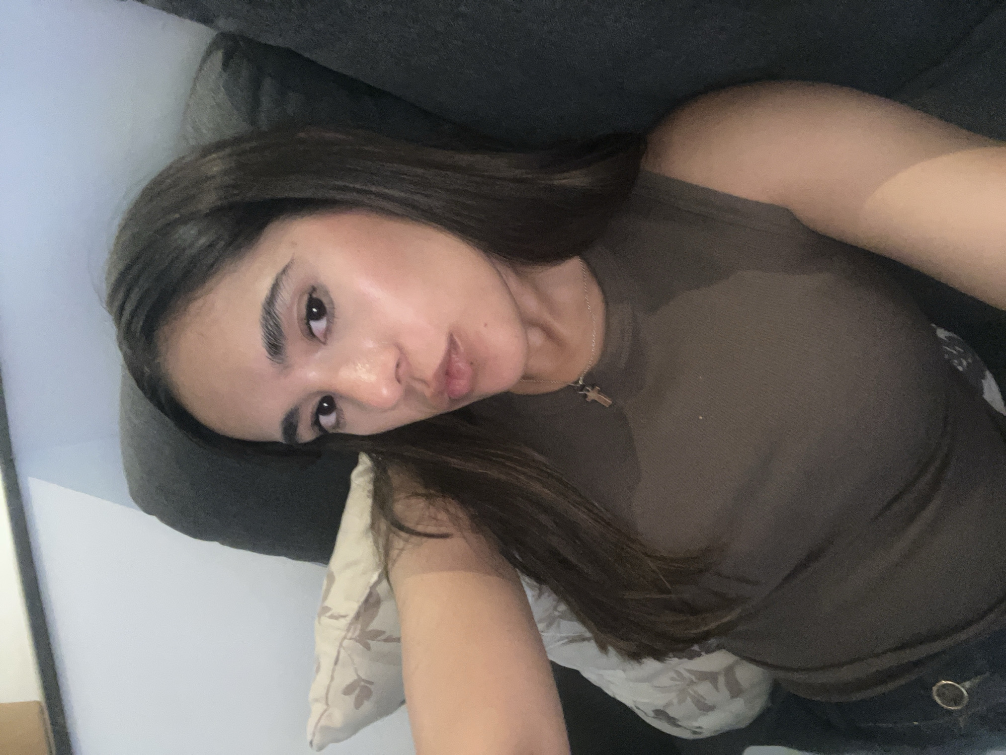 A young woman with long, dark brown hair is reclining on a gray sofa. She is wearing a brown tank top and a silver necklace with a cross pendant. A patterned pillow rests beneath her head. She is looking directly at the camera. The location is unknown.