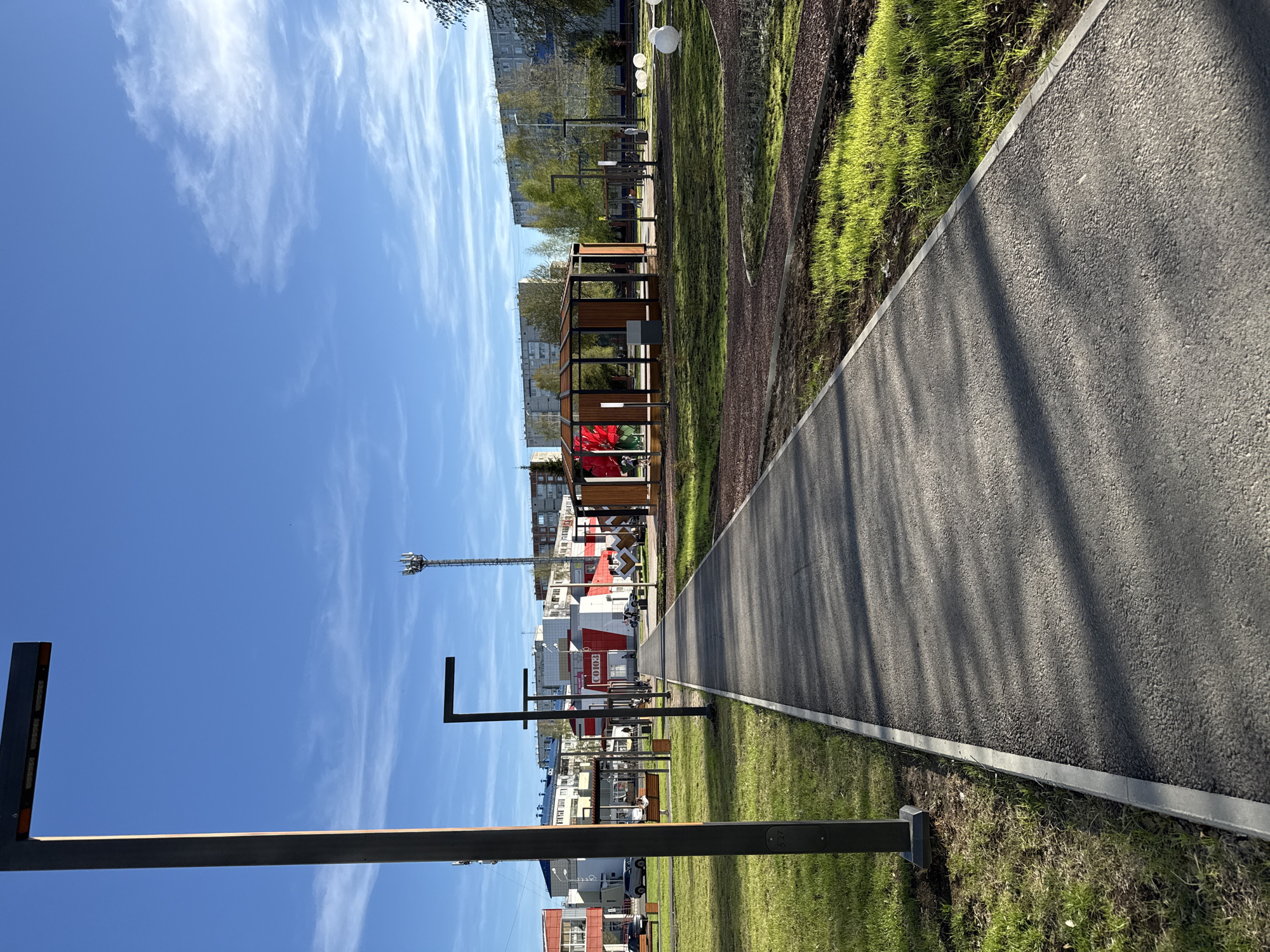 This is a photograph taken in Tobolsk, Russia. A straight, gray asphalt walkway stretches into the distance, its surface textured and lined with shadows, bordered by a strip of green grass on one side and a darker mulched area on the other. Beyond the walkway, modern architectural structures are visible including a cell phone tower, buildings and a wooden pavilion. The buildings include a building with the word "Союз" on it. The scene is bathed in sunlight under a bright blue sky dotted with wispy white clouds. In the foreground are dark metal structures, likely lampposts or decorative elements lining the path.