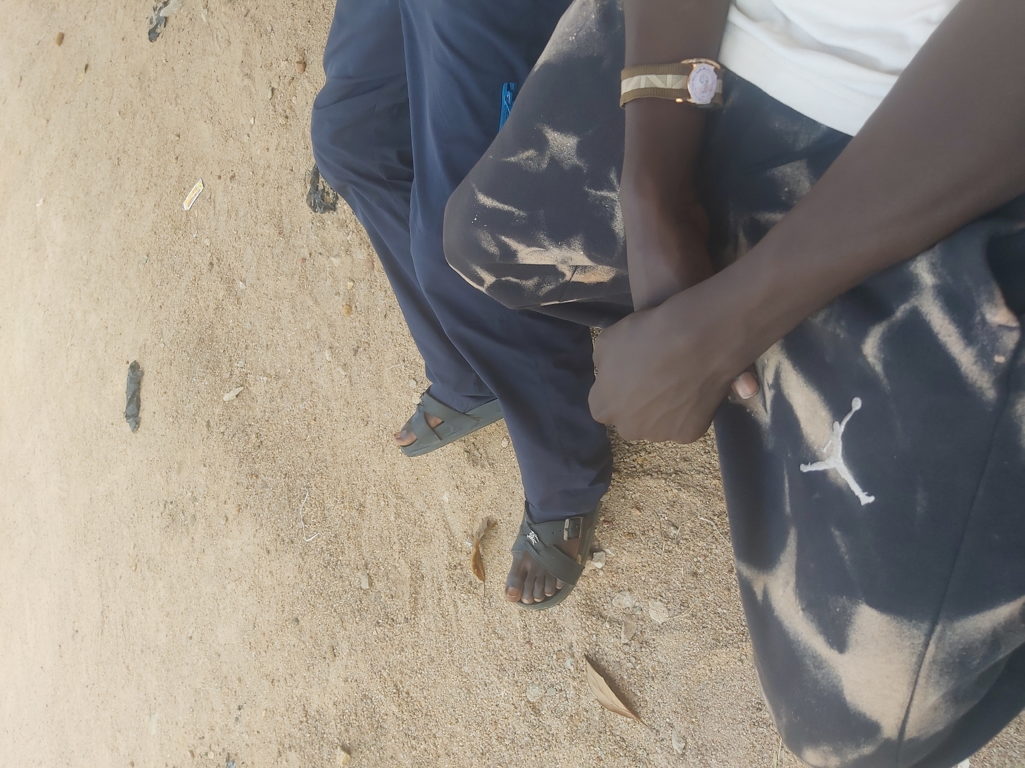 The image depicts a close-up view of two individuals resting on sandy ground in Jos, Nigeria. The foreground shows the legs and arms of two people. The left person is wearing dark pants with a tie-dye pattern and a white t-shirt. A watch with a light-colored band is visible on their right wrist. The right person is wearing dark blue trousers and dark sandals. The ground is composed of sand and small pebbles, with a few dry leaves scattered around. In the background, there are a few pieces of dark debris. The scene suggests a relaxed outdoor setting.