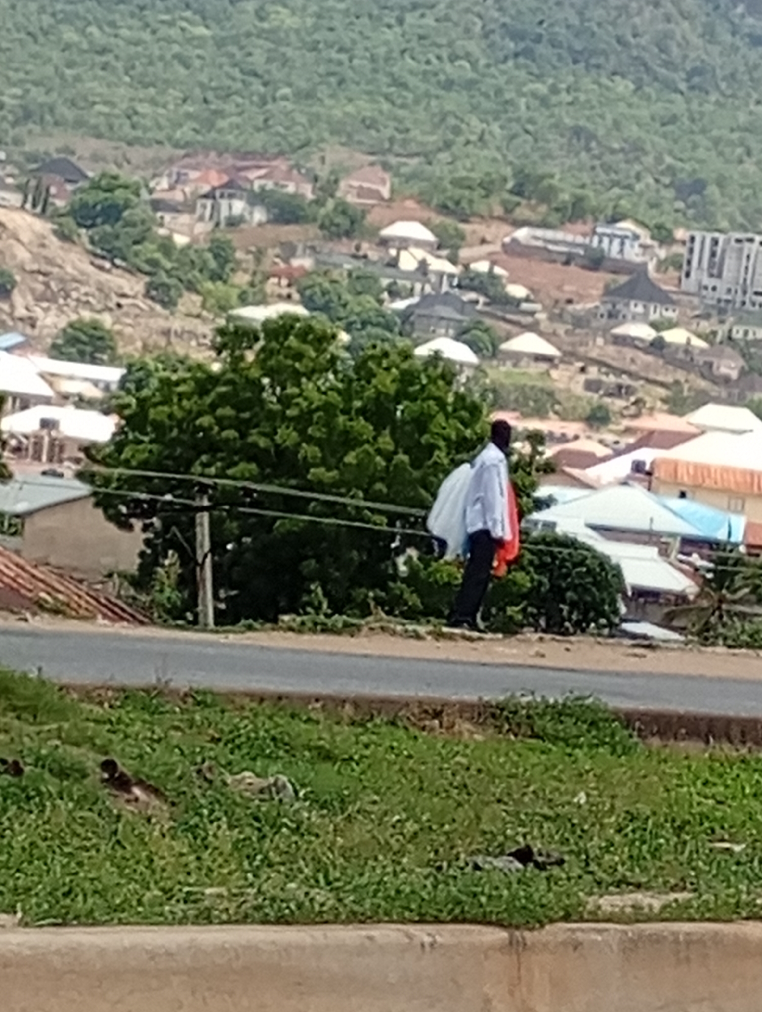 A medium shot captures a man walking along a roadside in an urban area of Unknown, Unknown. The man, dressed in dark trousers and a light-colored shirt, carries bags of white and red material. The background reveals a landscape of low-lying buildings interspersed with trees. A concrete road runs horizontally through the center of the image, with a grassy embankment in the foreground. Above the road, buildings are densely packed on the hills. The sky is overcast, contributing to the soft lighting of the scene.