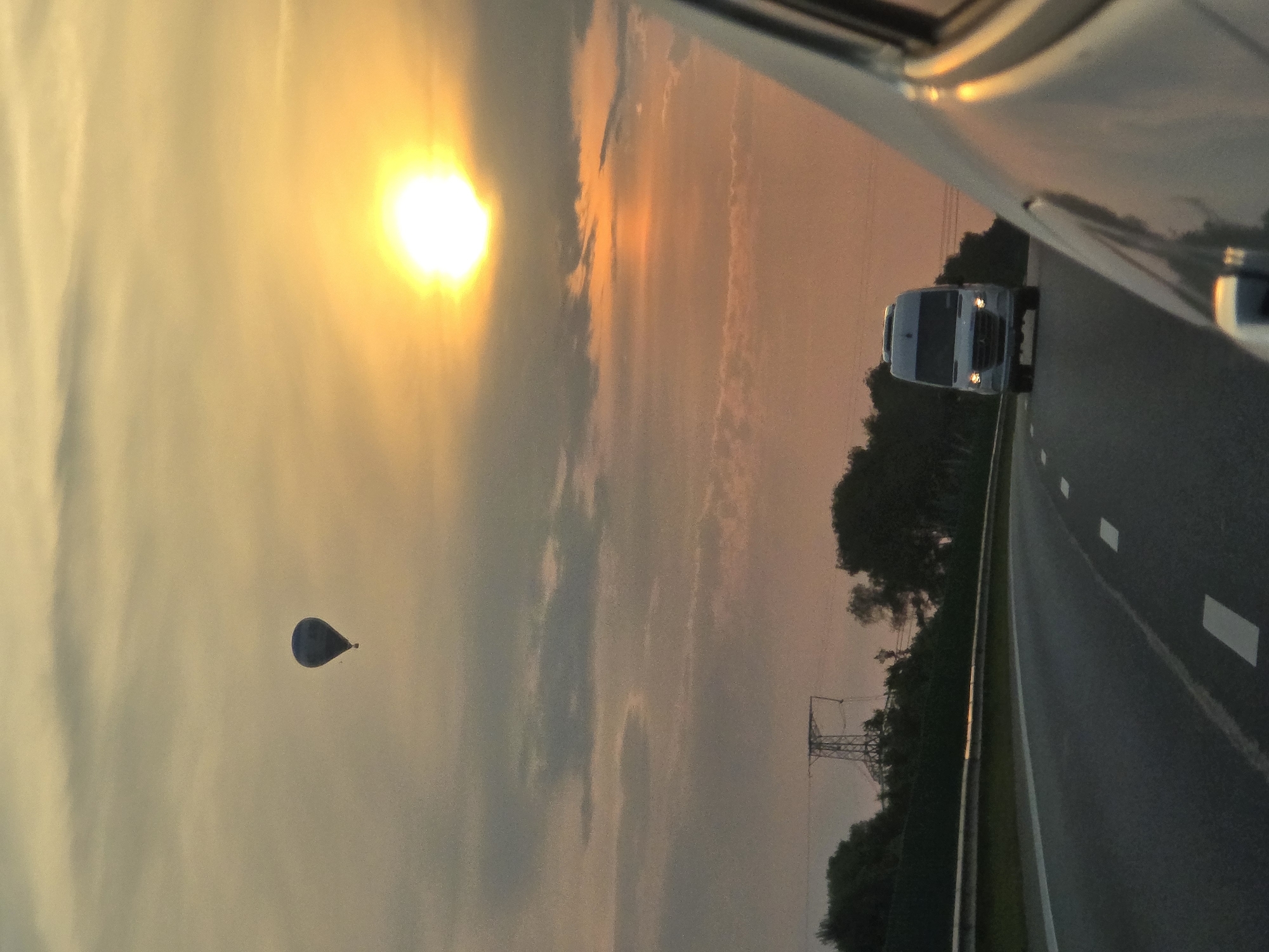 The image captures a sunset scene on a highway in Lazdėnai, Lithuania. A hot air balloon is visible in the sky, floating above a brightly lit sun. In the foreground, a road with white lane markings curves to the right. A white van is driving in the same direction as the viewer, in front of another vehicle, which is partially visible on the right side of the frame. Trees and a power line tower can be seen on the left side of the highway. The sky is filled with clouds, tinged with orange and yellow hues from the setting sun.