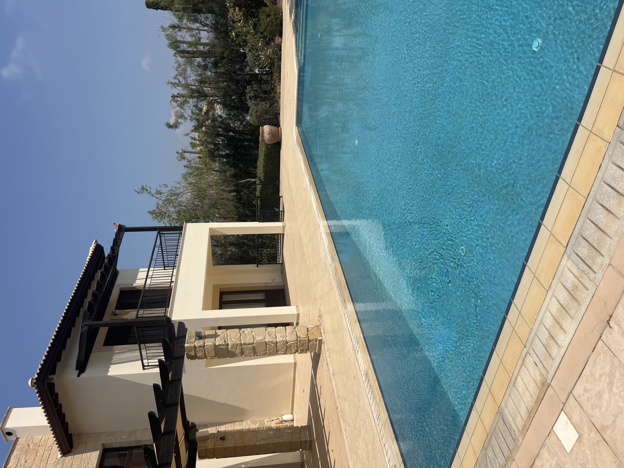 A bright, sunny outdoor scene showcases a large, clear blue swimming pool in the foreground, with textured light-colored tiles surrounding its edge. To the left and behind the pool, a cream-colored villa with a dark-tiled roof and a second-story balcony with a black railing is visible. The villa has stone pillars supporting a pergola that extends over the tiled patio area. Beyond the villa and pool, dense green foliage and trees form a natural backdrop under a clear blue sky. The location is identified as Aphrodite Hills, Cyprus. There are no people or animals present in the image.