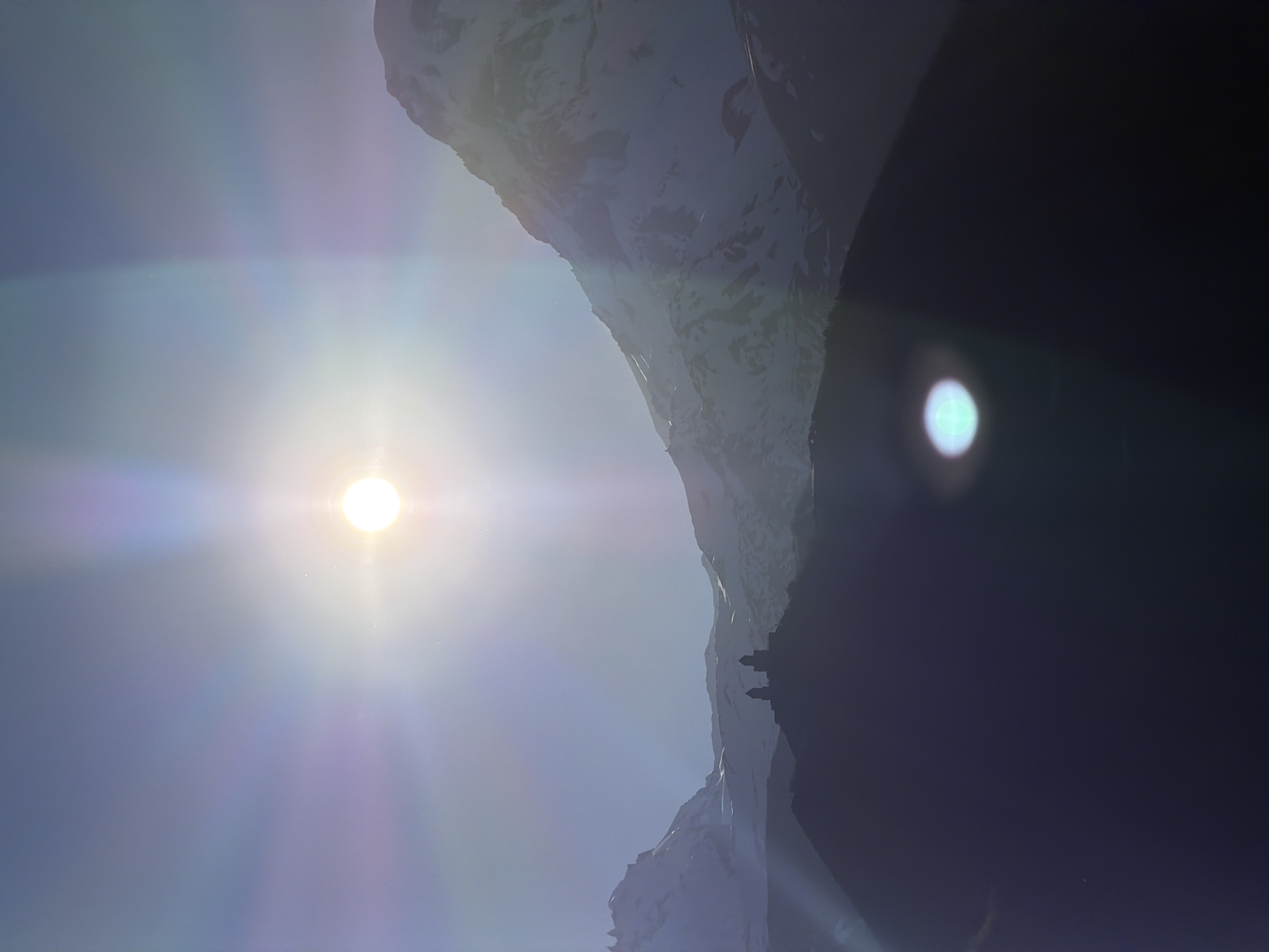 The image shows a bright sun in the sky with a strong lens flare. To the right of the sun, a mountain range is visible, partially covered in snow. The right side of the image is dark, obscuring the details in that area. There is a structure on the mountain, possibly the Gergeti Trinity Church, a popular landmark in Stepantsminda, Georgia. The sky is a clear blue, but the glare of the sun makes it difficult to see clearly.