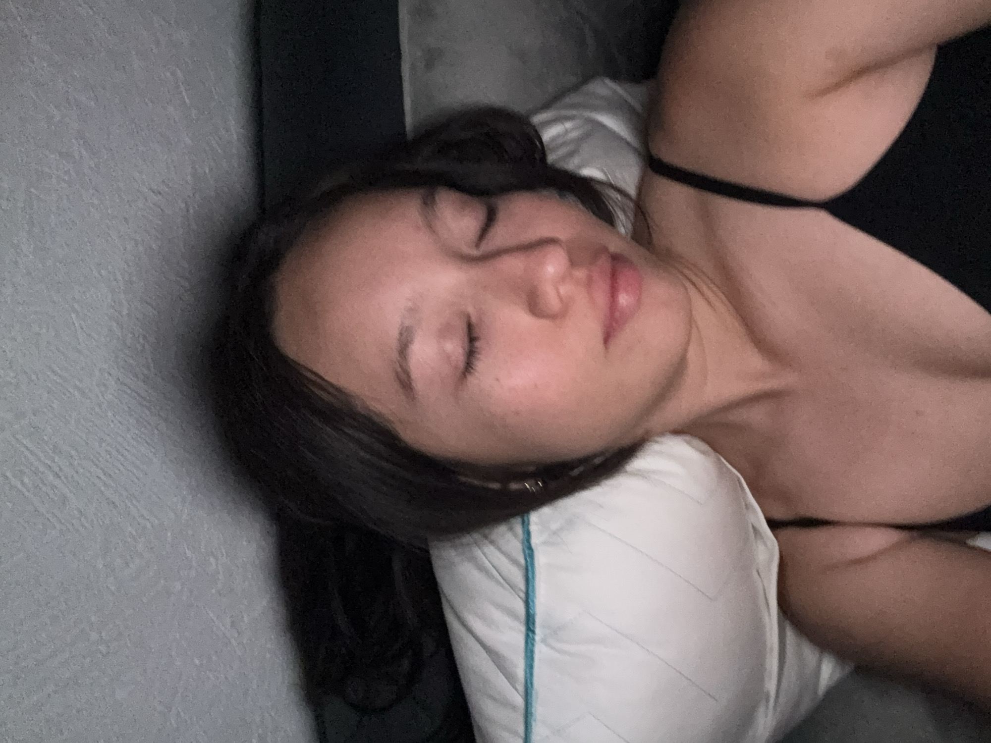A woman lies on her side on a bed with a white pillow with a teal border. She is wearing a black top and is facing the camera. The background consists of a gray wall. The location is unknown and there is no information to suggest it is in Puebla, Mexico.