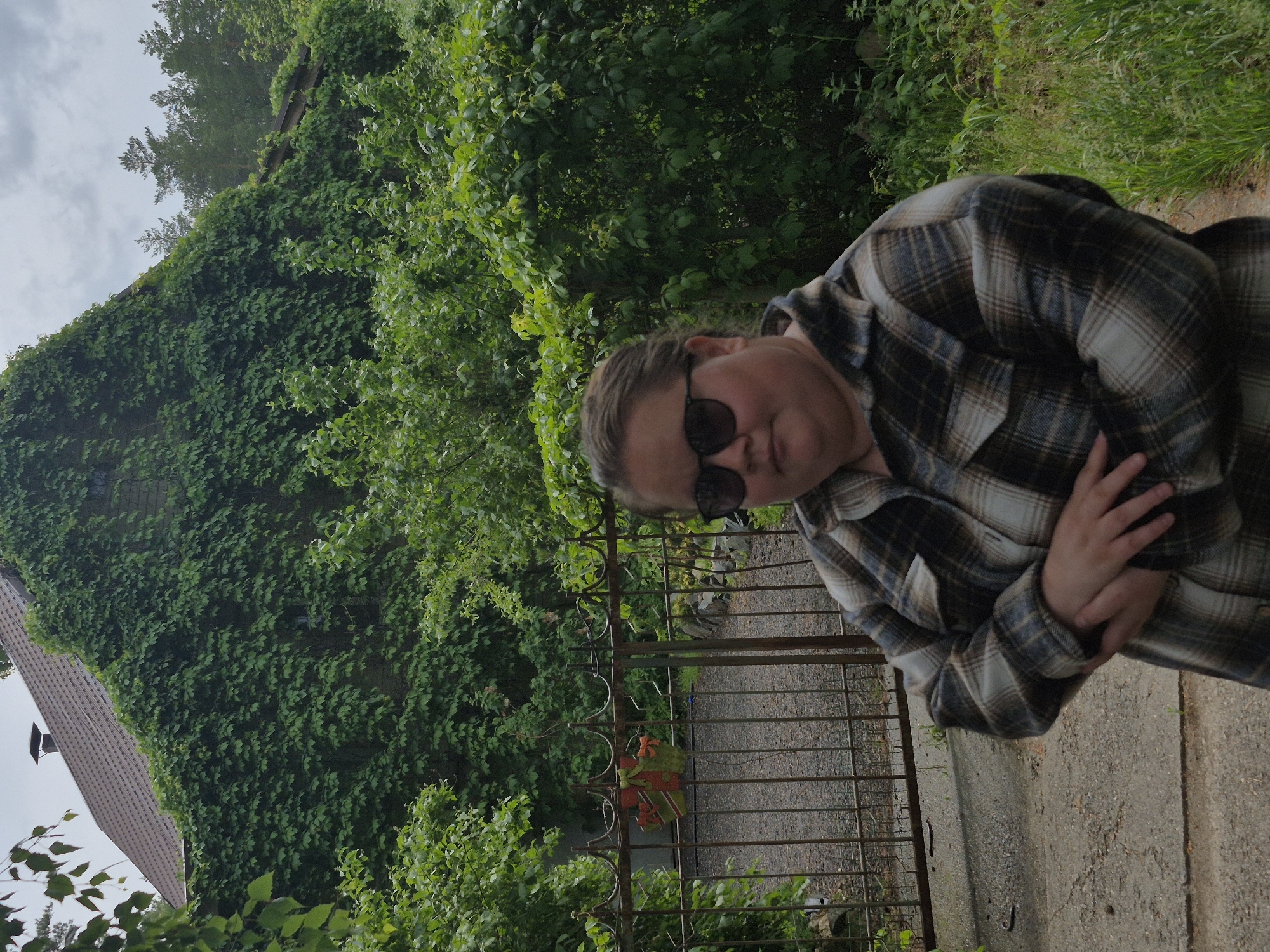 The image shows a person, likely female, from the chest up, positioned on the right side of the frame. They are wearing a plaid shirt with shades of brown, black, and cream, and dark sunglasses. Their arms are crossed, and light brown hair is pulled back.
Behind the person, to their left, is a rustic metal gate with decorative elements, possibly showing signs of rust. A small, multi-colored ornament, appearing to be fabric in red, green, and orange, hangs on the gate. A paved or concrete path extends behind the person and through the gate area, with patches of green grass and bushes visible beyond it.
In the background, predominantly to the left, is a house featuring a reddish-brown tiled roof and a chimney. The visible wall of the house is almost entirely covered in dense, vibrant green ivy or climbing plants. Tall green trees rise above the house, silhouetted against an overcast, grey sky. The overall setting is lush with greenery, suggesting a natural or overgrown outdoor environment.
The time of day appears to be daytime, with an overcast sky providing soft, diffused light. The location is consistent with a rural or garden setting in Noviy, Russia. No visible text is present in the image.
