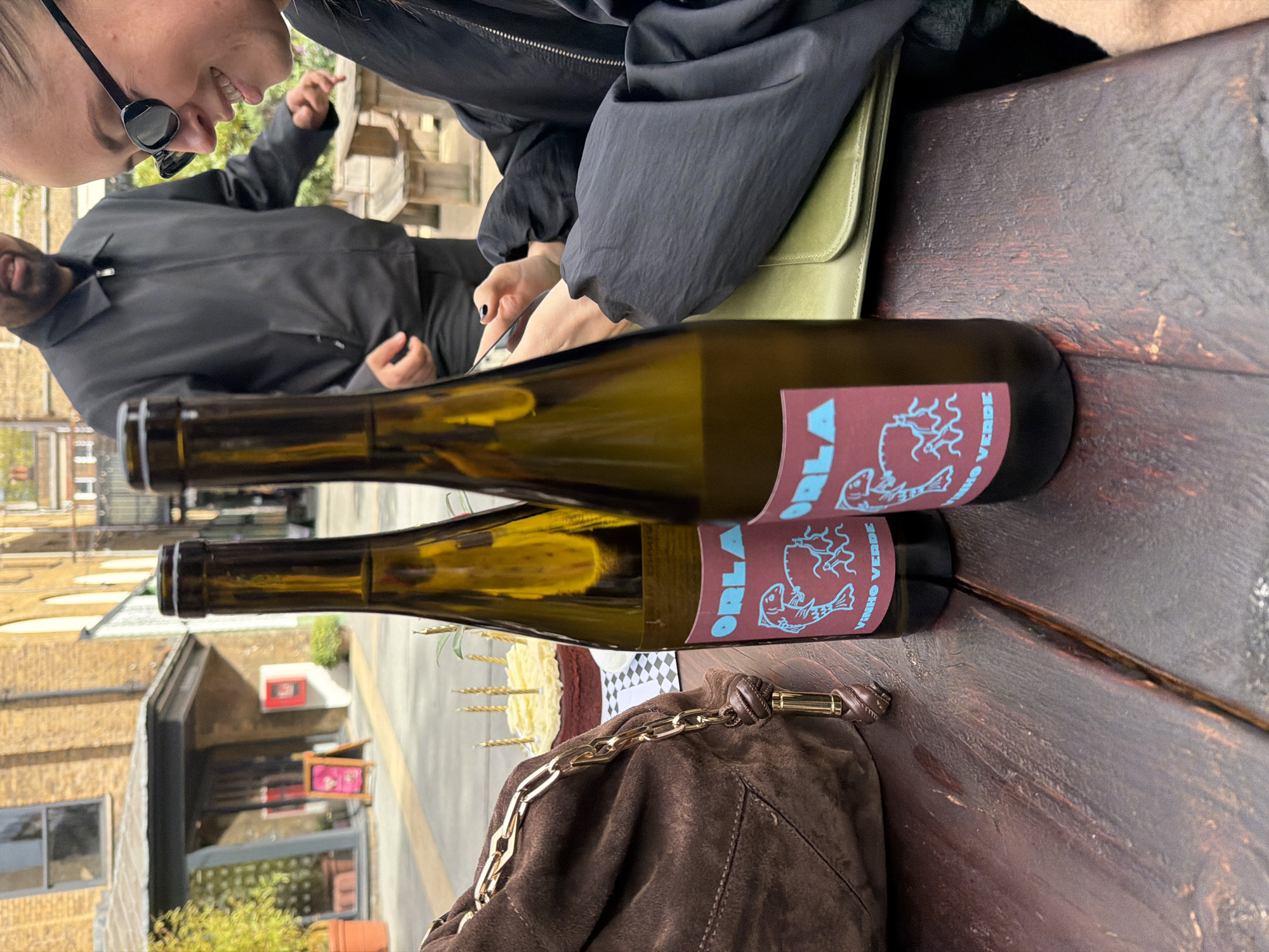 Two bottles of "ORLA Vinho Verde" wine are on a dark brown wooden table. One bottle is stacked partially on top of the other, obscuring the label of the bottom bottle. A brown suede purse with a thick gold chain is near the bottom of the frame. A partially visible white cake with gold candles is next to the purse. Two people are visible in the upper left, one wearing sunglasses and smiling. They appear to be dining or socializing outdoors. The location appears to be in London, United Kingdom due to the style of the architecture in the background.