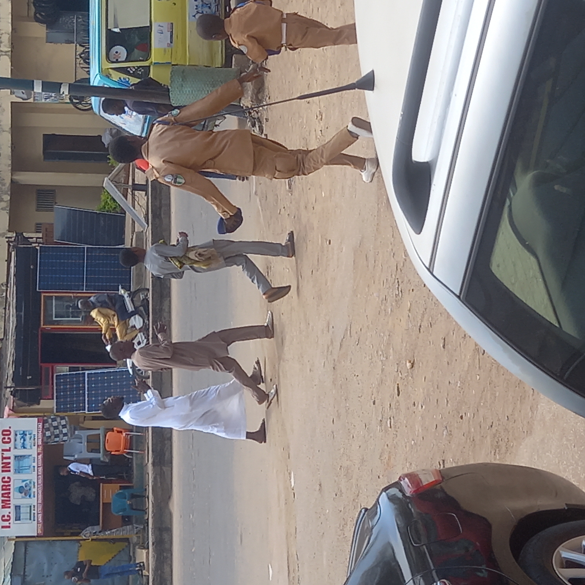 The image shows a street scene in Dadi Riba, Nigeria. In the foreground, a portion of a white vehicle and a black vehicle are visible. Several people are walking on the side of the road. They are dressed in casual and traditional clothing. One person is wearing a brown uniform with a white stripe on the pants, and holding a long black object. Another individual is wearing a long white robe, with the other wearing shades of gray. The setting includes shops, solar panels, and a yellow vehicle in the background. A sign for "I.C. MARC INTL CO." is visible on one of the buildings.