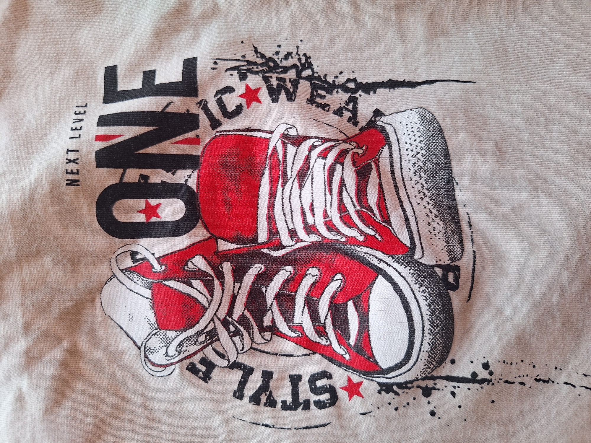 The image displays a close-up, overhead view of a graphic print on a beige t-shirt. The graphic features two red high-top sneakers with white laces, positioned diagonally across the fabric. Above the sneakers, in large black block letters, is the word "ONE". Above "ONE", in smaller black text, are the words "NEXT LEVEL". To the left of the sneakers, vertically oriented, is the word "STYLE" in black. To the right of the sneakers, also vertically oriented, is the word "MAGIC" in black. Red stars are interspersed within the text and the graphic. Splatter and streak elements in black ink surround the sneakers, suggesting a dynamic, street-art aesthetic. The fabric of the t-shirt shows visible knit texture. There are no people or any identifiable elements of the city of Varna, Bulgaria, visible in this image.