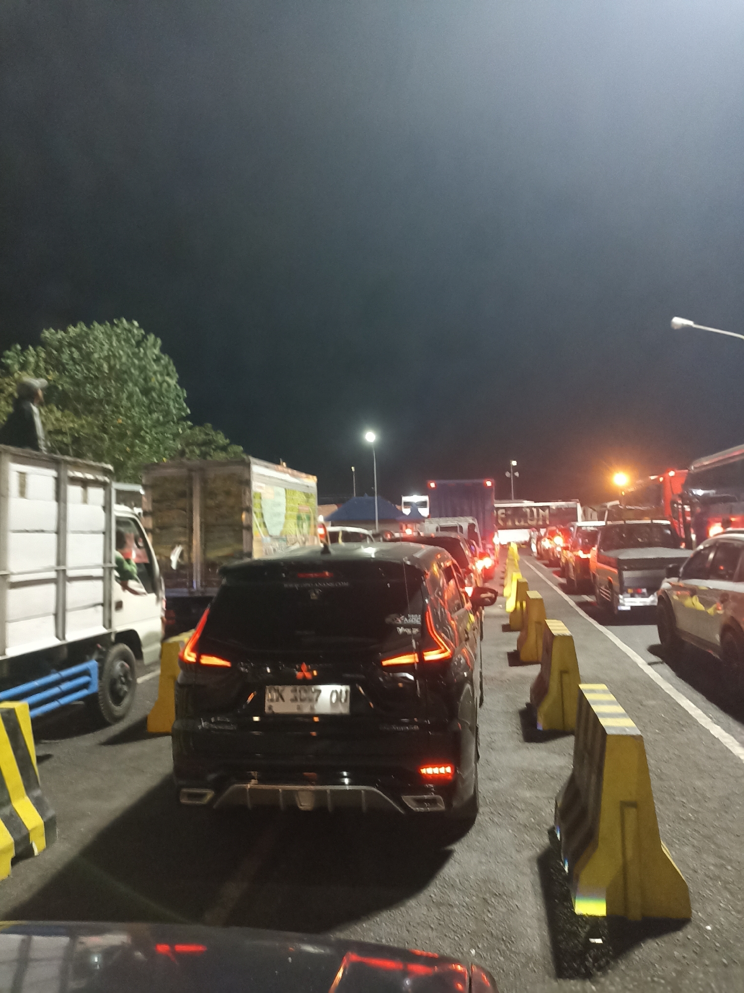 The image depicts a traffic jam at night in Melaya, Indonesia. A black car, a white truck, and several other vehicles are visible, stopped on an asphalt road. The road is lined with yellow and black traffic barriers. The sky is dark, suggesting nighttime, and illuminated by streetlights and vehicle headlights. There are no discernible individuals or specific actions occurring beyond the presence of vehicles in a standstill. The environment appears to be an outdoor road or highway.