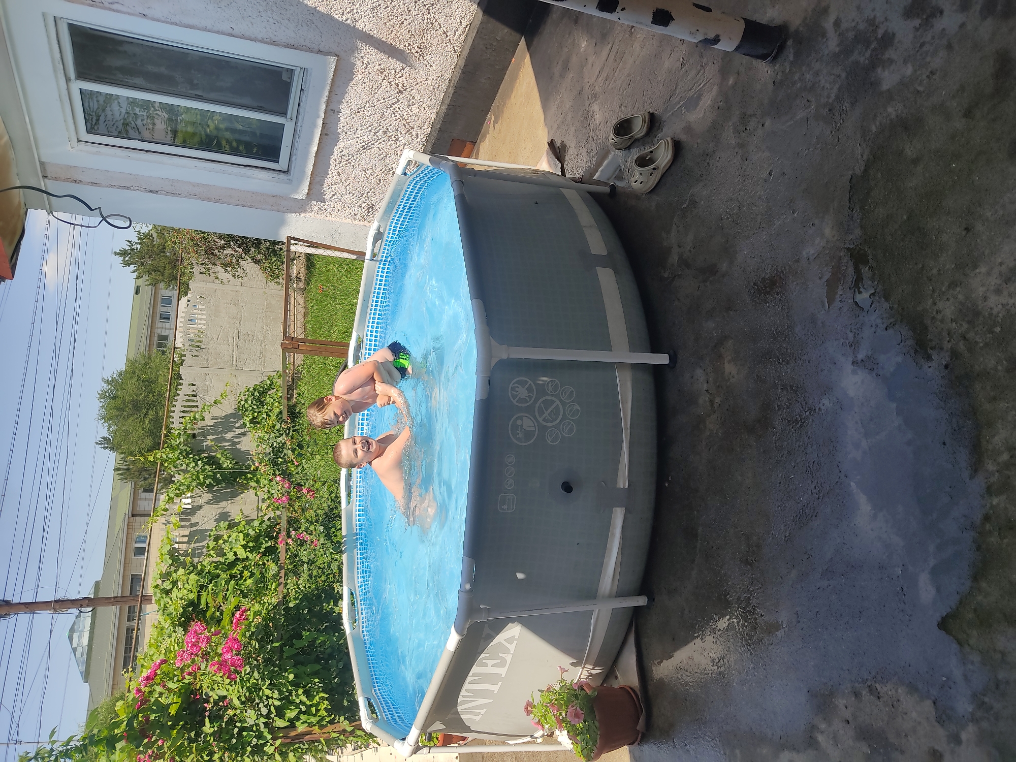 The image depicts two young boys playing in a round, above-ground swimming pool filled with clear blue water. The pool is situated on a dark, wet concrete patio, likely in a residential backyard. Green foliage and pink flowers are visible in the background, as is a pale, textured wall of a building with a white-framed window. A pair of light-colored clogs rests on the patio near the pool. The scene appears to be outdoors on a sunny day. The presence of an "INTEX" logo on the side of the pool suggests it is a commercially manufactured product. The setting is a backyard in Shamalgan, Kazakhstan.