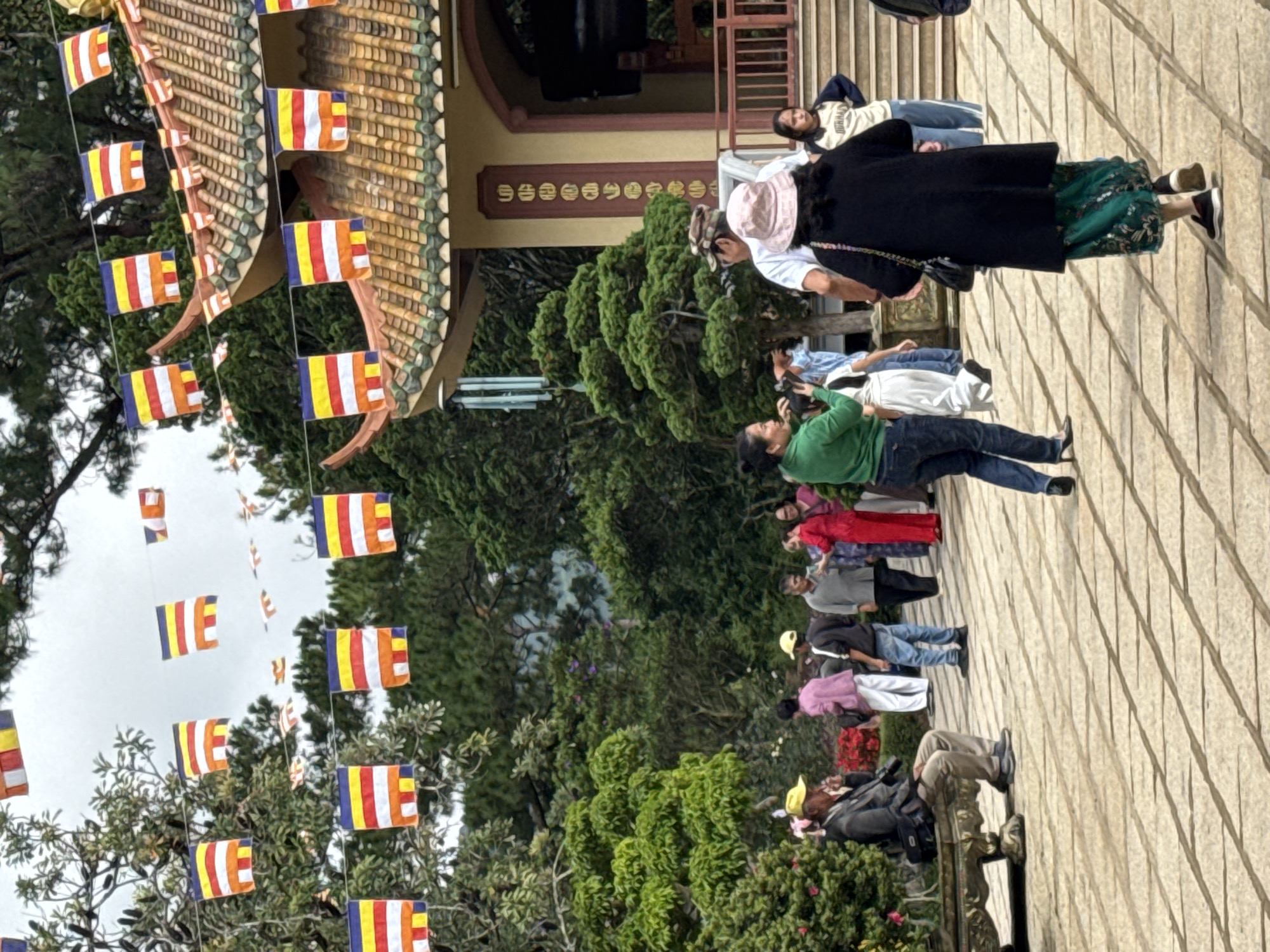 This image depicts an outdoor scene at a Buddhist temple in Da Lat, Vietnam. The paved foreground consists of light-colored rectangular stone tiles with dark grout lines arranged in a grid pattern. In the left foreground, a man wearing a yellow cap, dark jacket, light trousers, and dark shoes sits on a ornate stone bench. He holds a dark object, likely a camera, and a dark shoulder bag rests beside him on the bench. Behind him are manicured green bushes, some with small red flowers. In the central foreground, a woman with dark hair, wearing a green long-sleeved top and blue jeans, stands facing left with her right arm raised, appearing to interact with someone off-frame. To her right and further back, another woman, wearing a black long coat, a patterned green and gold skirt, a light pink bucket hat, and dark sneakers, walks away from the camera towards the right. A dark shoulder bag with a chain strap hangs on her left side. Numerous other individuals are visible throughout the midground, walking and standing. Notable figures include: * A man in a white t-shirt and camouflage cap, partially obscured, standing in the mid-right. * A woman in a white dress walking towards the right. * A man in a light grey collared shirt and dark trousers interacting with a woman in a red traditional dress. * A young girl in a beige and navy long-sleeved top with "DURHAM" visible on it, standing near the temple steps on the right. * A man in a black long-sleeved top and blue jeans stands to the left of the man in grey, facing right. The background features a multi-tiered temple building with curved, tiled roofs. The roof tiles are predominantly shades of orange-brown and green. A tall, dark brown vertical pillar adorned with golden Vietnamese or Chinese characters is visible on the temple's facade. To the right of the pillar, a large, dark, arched opening, possibly an entrance or housing a bell, is present. Brown wooden railings lead up to this entrance. A metallic wind chime hangs to the left of the temple building. The entire scene is set amidst lush green trees and foliage, some with visible grey branches, suggesting a natural and well-maintained environment. Numerous Buddhist flags, characterized by five horizontal stripes (blue, yellow, red, white, and orange), are strung on lines across the sky, between trees, and around the