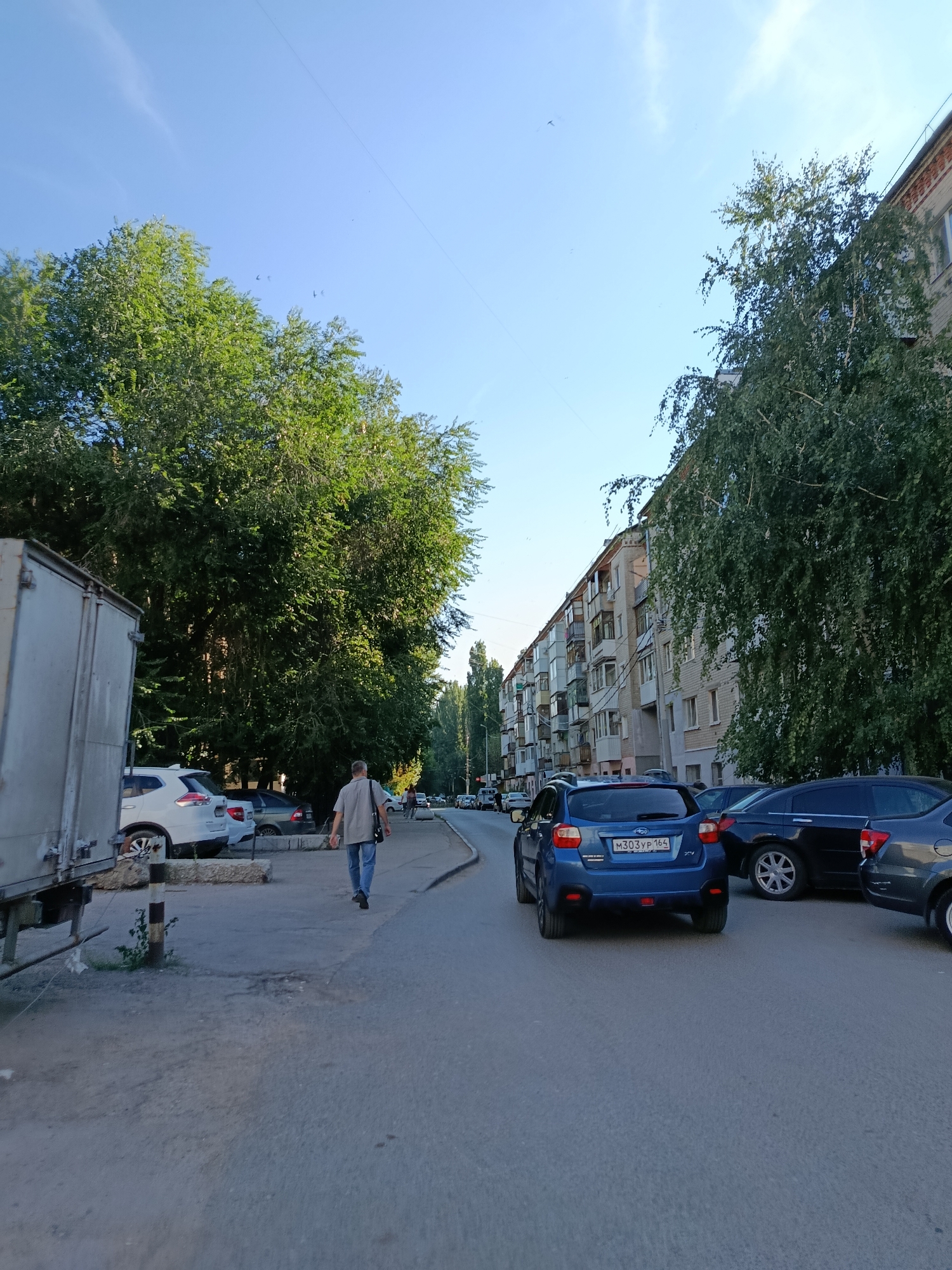 In Saratov, Russia, on a clear day, a man walks away from the camera down a paved street lined with parked cars and apartment buildings. The man, wearing a light-colored shirt and blue jeans, carries a dark bag over his shoulder. On the left, a large, white truck trailer is parked. Beyond the trailer and a row of parked cars, lush green trees dominate the left side of the scene. On the right, a multi-story apartment building runs along the street, its facade showing balconies and windows. Several vehicles are parked and in motion on the street, including a blue Subaru. The sky above is a bright, clear blue.