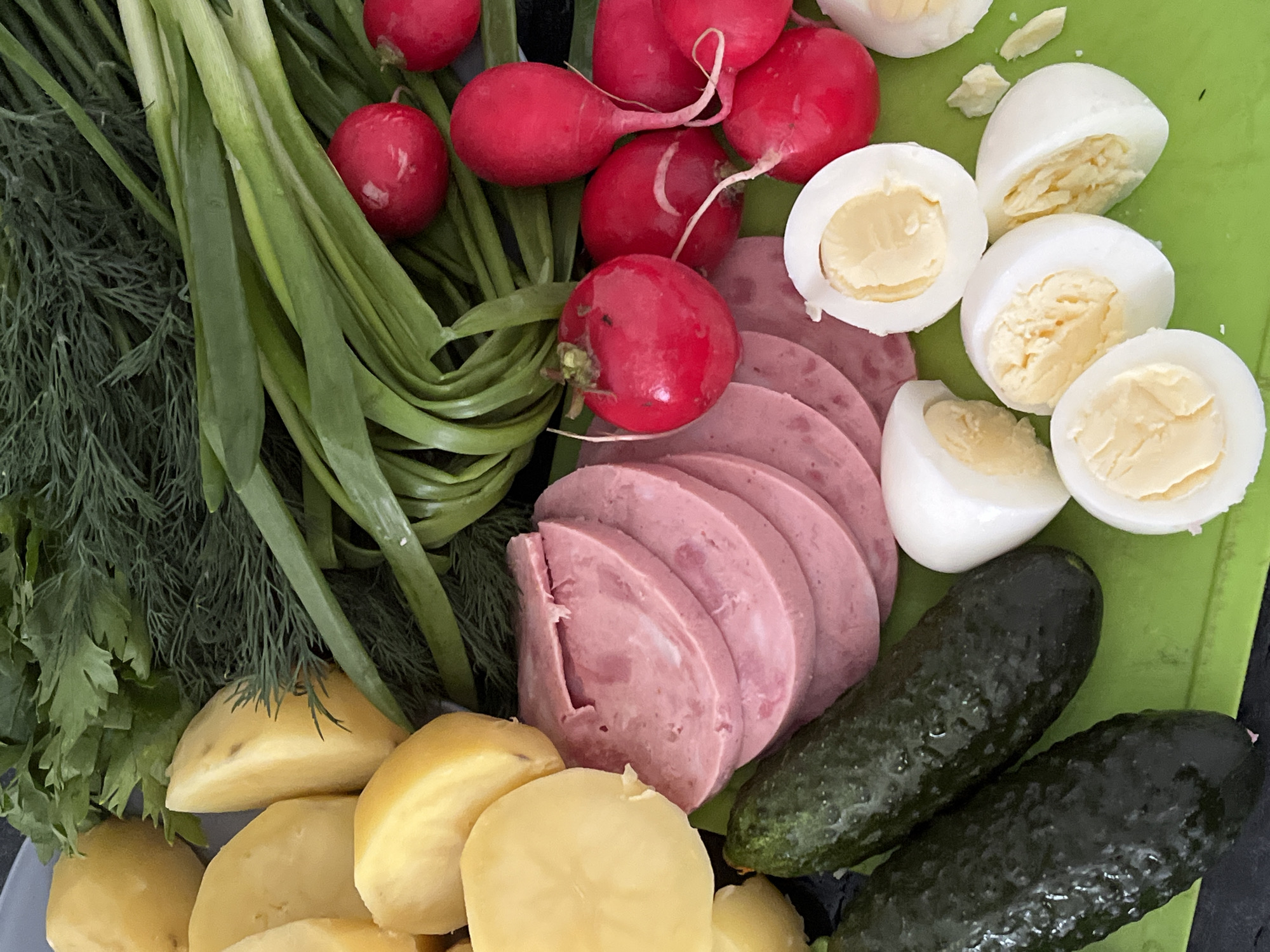 A close-up shot presents a selection of food items, likely ingredients for a dish, arranged on what appears to be a green cutting board. The assortment includes fresh green onions, dill, vibrant red radishes, slices of boiled sausage, halved hard-boiled eggs, boiled potatoes, and two dark green pickles. The components are densely packed, filling the frame, with the green cutting board only partially visible. No people or locations are identifiable within the image. The overall impression is one of fresh, unprepared ingredients ready for use.