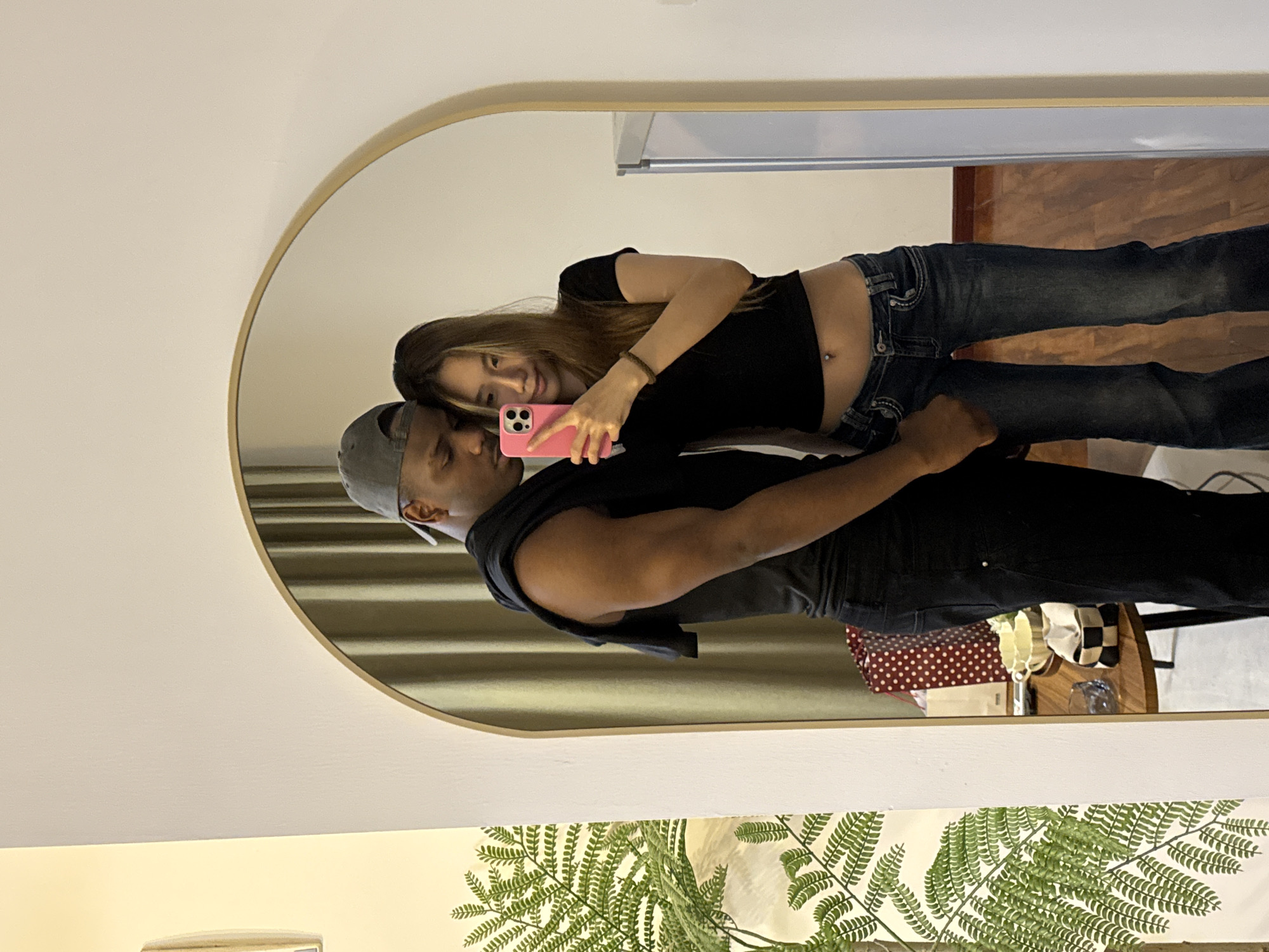 A couple is reflected in an oval mirror, taking a selfie with a pink phone. The woman has long brown hair, a black shirt, and jeans. The man wears a gray baseball cap and a black top. The background shows a room interior with curtains, a table with decorative items, and a glimpse of a window and hardwood floors. The location is unknown and cannot be determined as Yuzhong District, China from this image.