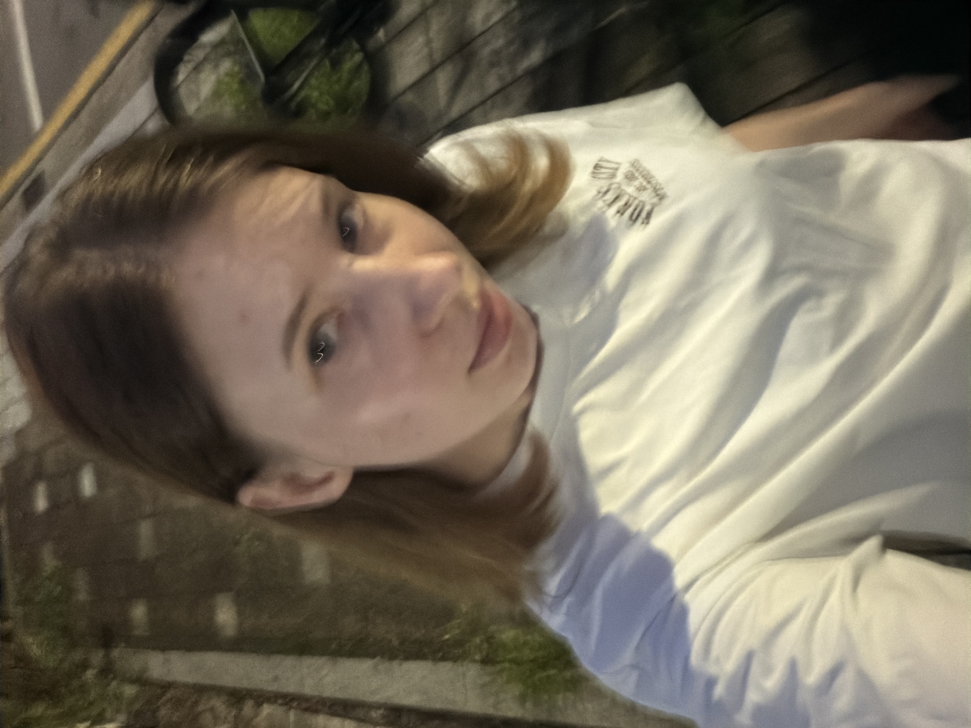 A young woman is taking a selfie outdoors at night. She is wearing a white t-shirt with a graphic on the chest. The background is blurred, but appears to be an urban or park setting with some vegetation and paving. The woman has light brown hair and light-colored eyes. She is looking directly at the camera and has a slight smile. The lighting suggests artificial light sources, possibly streetlights or building lights. The image quality is somewhat grainy due to low light conditions. No specific landmarks of Dongducheon-si are identifiable.