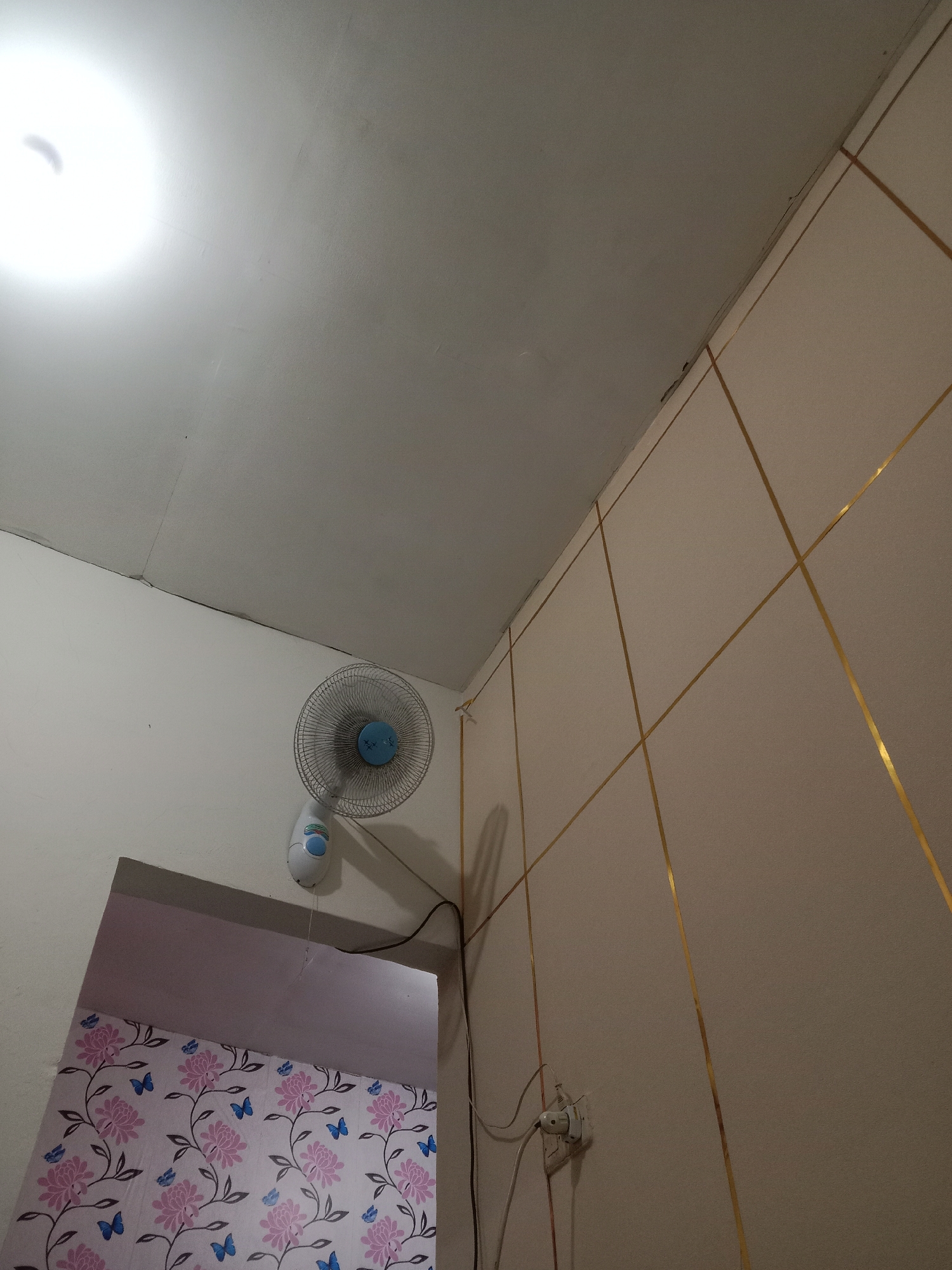 The image captures an interior scene, likely a room in Brangsong, Indonesia. In the upper left, a bright light source illuminates a textured ceiling with visible cracks and imperfections. Below the ceiling, a white wall extends downwards, meeting a section of wall adorned with a pink floral wallpaper featuring blue butterflies. Mounted on the white wall is a white oscillating fan with a blue center, its power cord extending downwards. To the right, the wall transitions to a tiled surface with a pattern of intersecting gold lines. A white electrical outlet with a plug and wires is visible on this tiled section. The perspective suggests the photo was taken from a low angle, looking upwards towards the ceiling and walls.