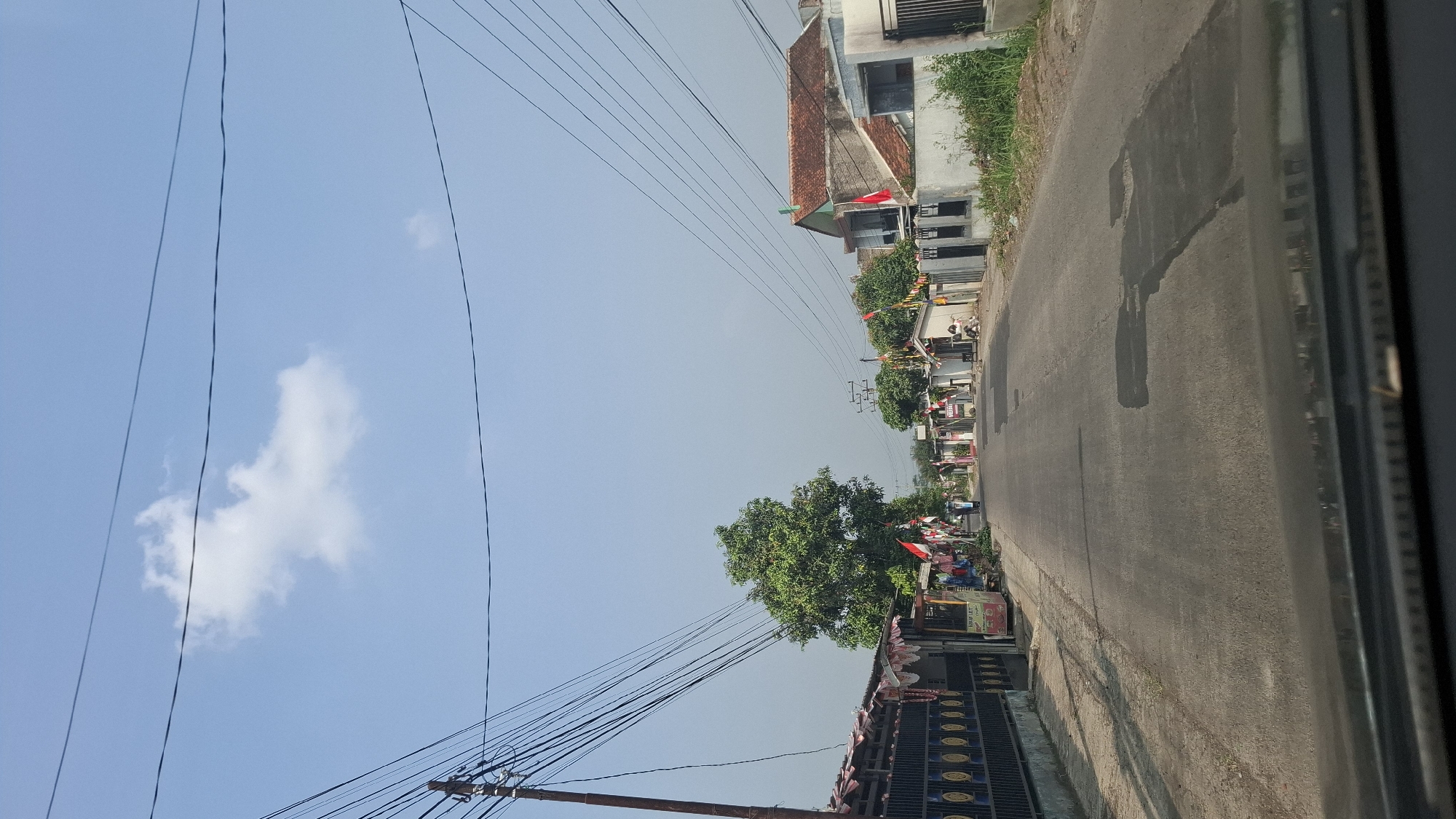 The image captures a street scene in Sukasari, Indonesia, under a clear blue sky with a few scattered clouds. The perspective is from a vehicle, showing the road ahead lined with houses and shops on both sides. Indonesian flags are prominently displayed along the street, suggesting a celebration or national holiday.
On the left side of the road, a black metal fence encloses a property with decorative circular patterns. Adjacent to the fence are several small stalls or shops, some appearing to sell food or drinks, with merchandise displayed outside. A large, leafy green tree stands tall behind these stalls.
On the right side of the road, houses with red-tiled roofs are visible. Some of these houses have white walls and dark-framed windows. Small shops or buildings are also present, with flags and decorations. In the foreground on the right, overgrown green vegetation is seen.
The asphalt road itself has several patches and marks, indicating wear and tear. In the distance, a few people can be seen walking and a motorcycle is present, indicating normal daily activity. The overall impression is of a residential and commercial street decorated for a special occasion in a tropical environment.