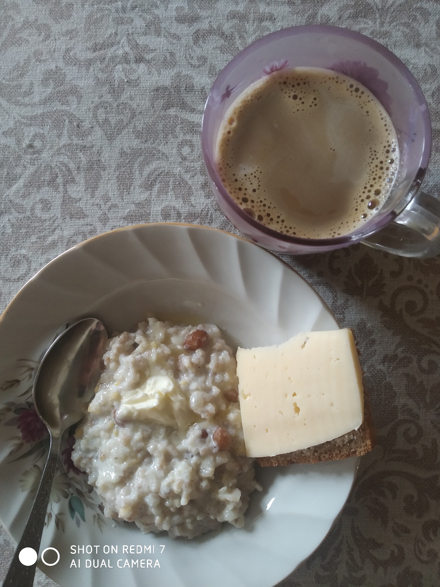 A high-angle shot depicts a meal consisting of porridge, cheese on bread, and coffee. The porridge, served in a white floral-patterned bowl with a golden rim, contains what appears to be butter and raisins. Next to it is a slice of yellow cheese atop a piece of dark bread. A spoon rests on the plate, beside the porridge. To the upper right, a transparent mug filled with coffee sits on the table. The table is covered with a neutral-toned, patterned tablecloth. The image was taken on a Redmi 7 phone with AI dual camera. The location is not discernible from the image.