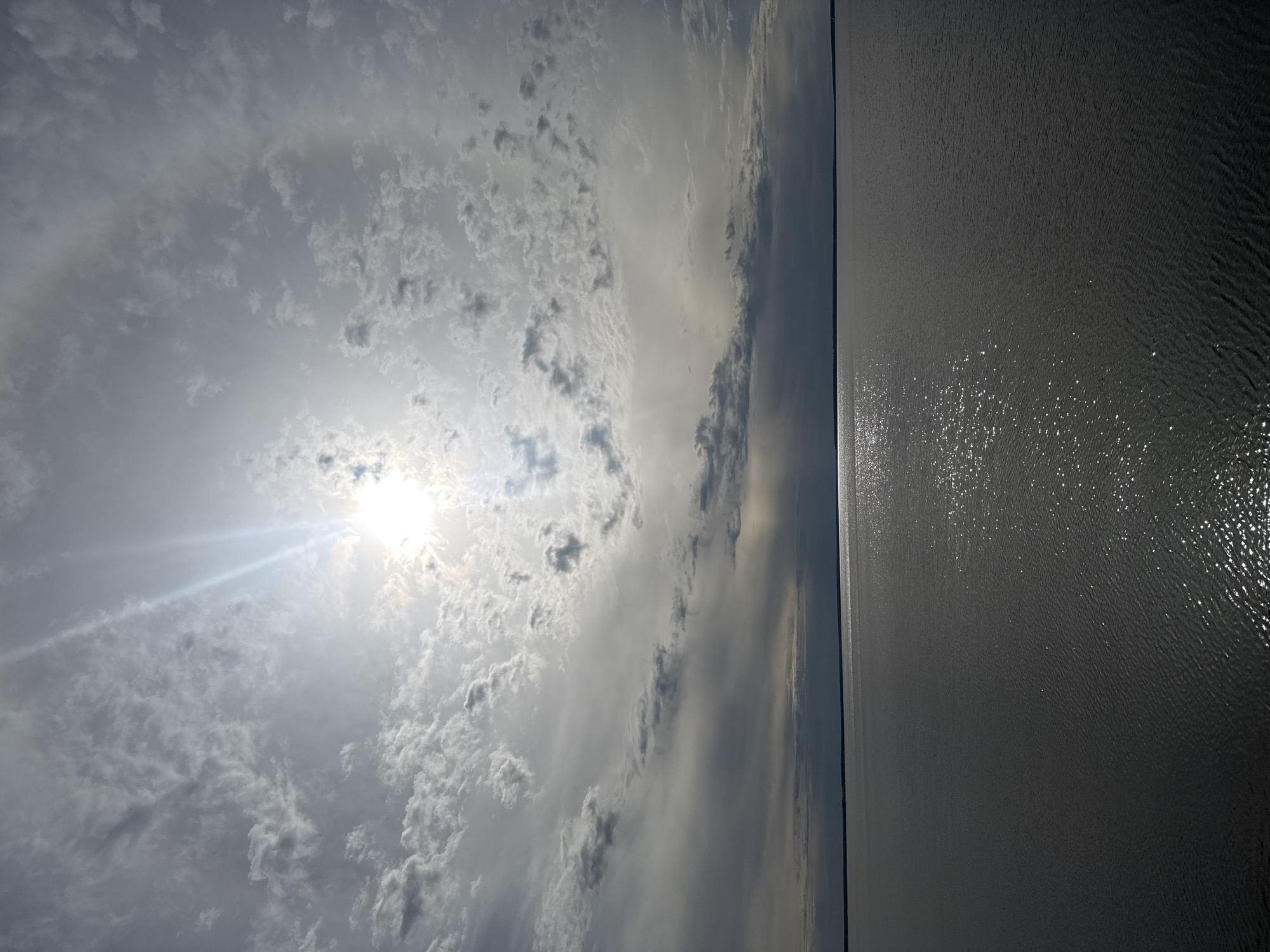 The image depicts a body of water, possibly a lake or large river, under a cloudy sky. The right side of the image shows the water, reflecting the sunlight with scattered bright spots. The water's surface appears rippled, suggesting a slight breeze. The left side of the image shows the sky, dominated by a mix of thin, scattered clouds and thicker, darker cloud formations. The sun is visible, partially obscured by the clouds, emitting bright rays.
Given the provided location information, this scene might depict the view of a nearby lake or river from the Timiryazevskii S/S area of Ivanovo, Russia. Without specific identifying features in the image, such as buildings or landmarks, it's impossible to pinpoint the exact location.