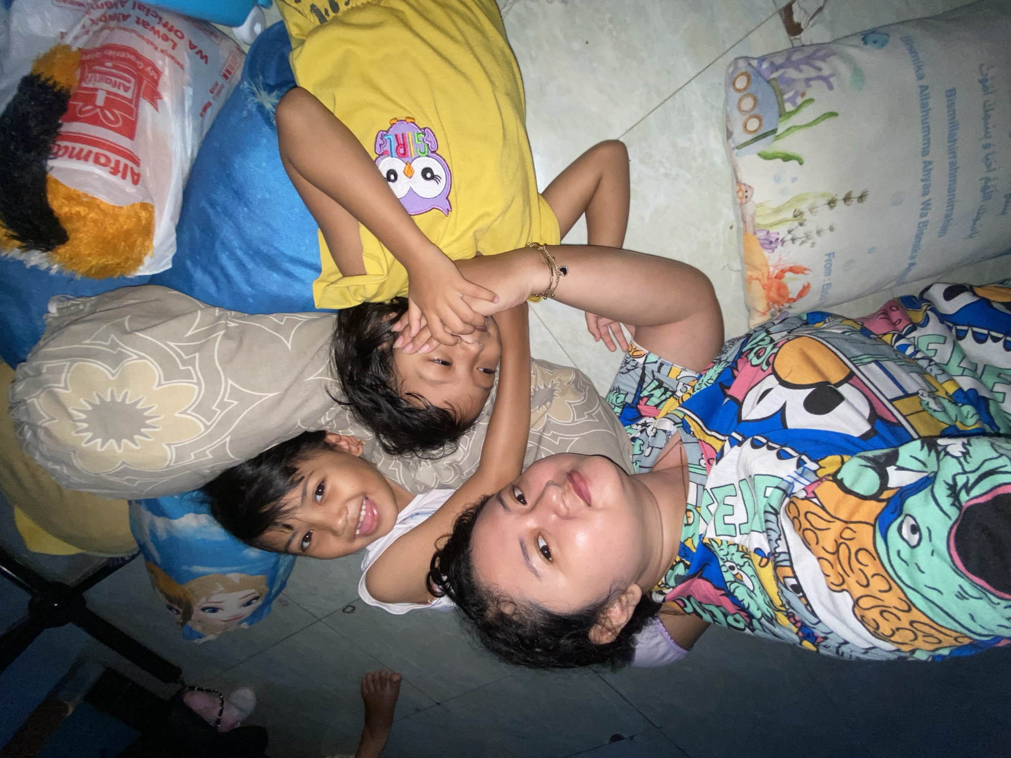 Three individuals are depicted lying on a light-colored tiled floor in an indoor setting in Gelam, Indonesia, viewed from above. On the right, an adult female with dark, curly hair and a round face lies on her back, wearing a multicolored, cartoon-patterned top and a visible gold chain. She looks directly towards the camera. To her left, a child with dark hair, wearing a yellow t-shirt with a purple owl graphic, lies on their back with hands clasped over their face; a gold-colored bracelet with a black charm is visible on their right wrist. To the left of this child, another child with dark hair, wearing a white sleeveless top, lies on their back, smiling broadly at the camera.
The environment includes several pillows: a solid blue pillow under the center child's head, a grey patterned pillow under the leftmost child's head, and a white pillow with an ocean-themed design (featuring a submarine, coral, and a red crab) and Arabic script visible to the right of the adult. A partially visible blue pillow depicting Disney's "Frozen" characters is located in the lower left. In the upper left quadrant of the image, a transparent Alfamart plastic shopping bag, containing orange and black fluffy material, rests on another blue pillow. The floor is light-colored tile with visible grout lines.