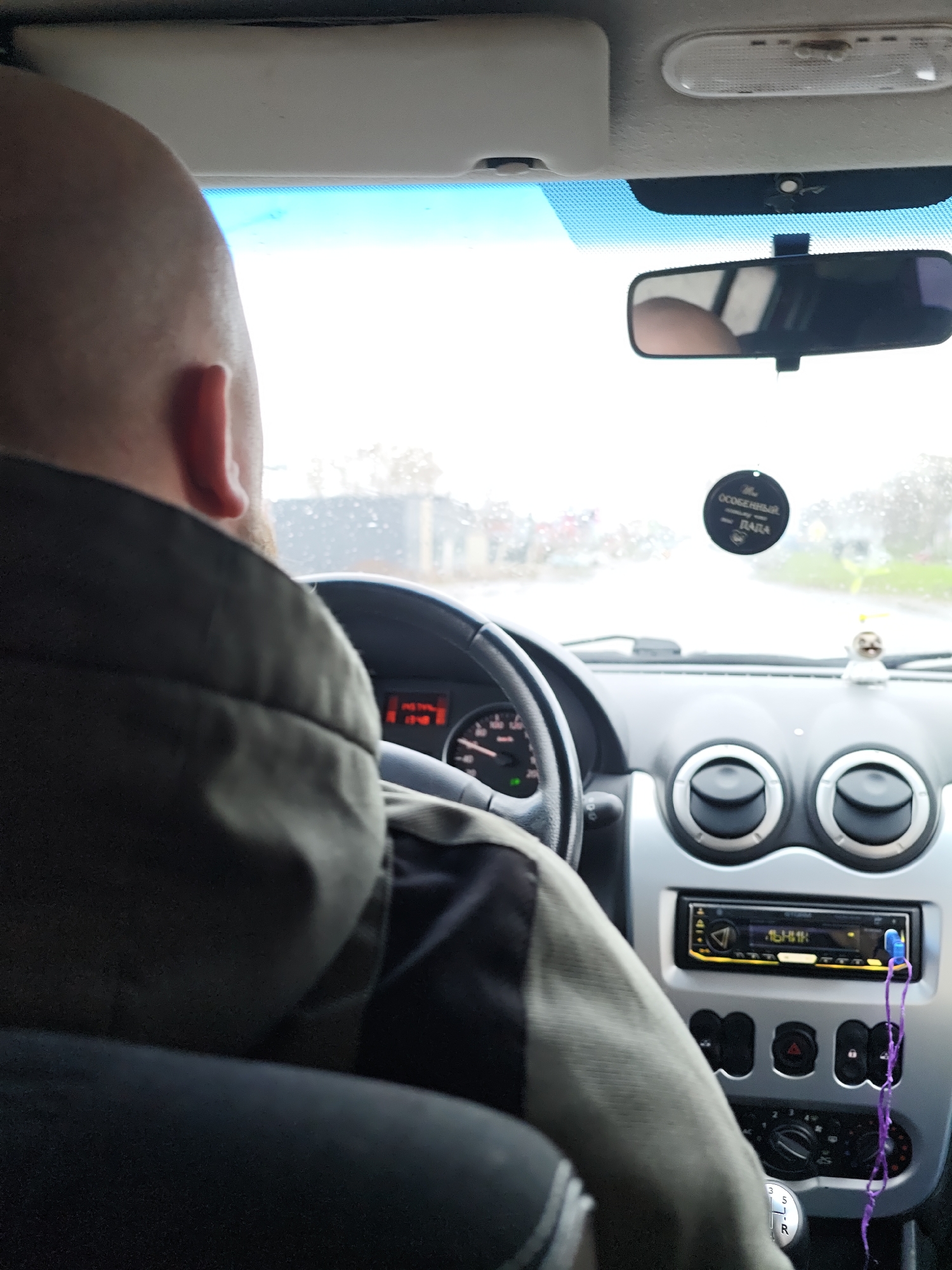 The image captures an interior view of a car from the back seat, looking forward at the driver and the road ahead. The primary subject is a bald man with a visible beard, seen from behind, wearing a dark green or khaki jacket, who is driving the vehicle. His hands are positioned on the steering wheel.
The car's interior features a dashboard with prominent circular air vents, a car radio with a blue display and a purple string or lanyard hanging from it, and a manual gear stick displaying "5 R 3 1". A speedometer, partially visible through the steering wheel, shows an unidentifiable low speed. On the passenger side of the dashboard, a small, light-colored figurine, possibly a sloth or animal toy, is placed. An oval-shaped, black air freshener hangs from the rearview mirror, which reflects a portion of the driver's head. The car's ceiling, including a sun visor and an overhead light panel, is also visible.
Through the windshield, the exterior scene appears overcast and slightly blurry, suggesting cloudy weather or recent rain, with visible moisture on the glass. A wet road stretches ahead, and indistinct buildings or trees are visible in the distance. The lighting is diffused, indicating daytime.
Visible text on the air freshener includes "ОСОБЕННЫЙ" (SPECIAL), "УЮТ" (COZY/COMFORT), and "ЛАДА" (LADA), which identifies the car brand. Some unidentifiable Cyrillic text is faintly visible on the car radio display. The scene depicts a routine moment of driving in what appears to be a Lada car, likely in Chaikovsky, Russia, given the context.