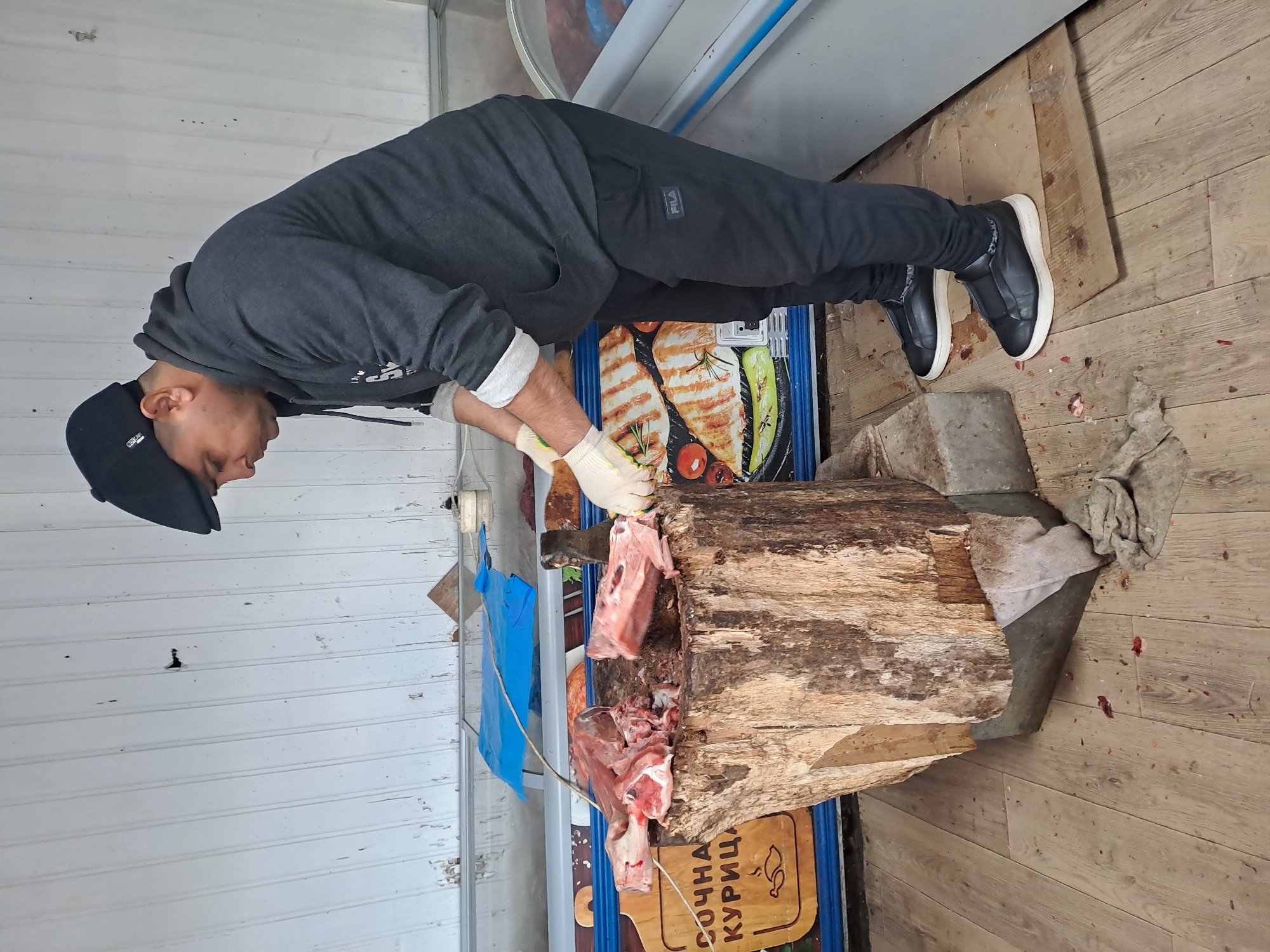 A man in Obukhovo, Russia, wearing a dark grey hoodie, black pants with a Fila logo, black shoes with white soles, and a black cap, is butchering meat on a large wooden block. He is wearing white gloves and using a cleaver. Pieces of meat are visible on the block and around its base. The background includes a cooler or display case, a wall with white paneling, and a floor with wood-patterned laminate. A sign in Cyrillic script is visible at the base of the block.