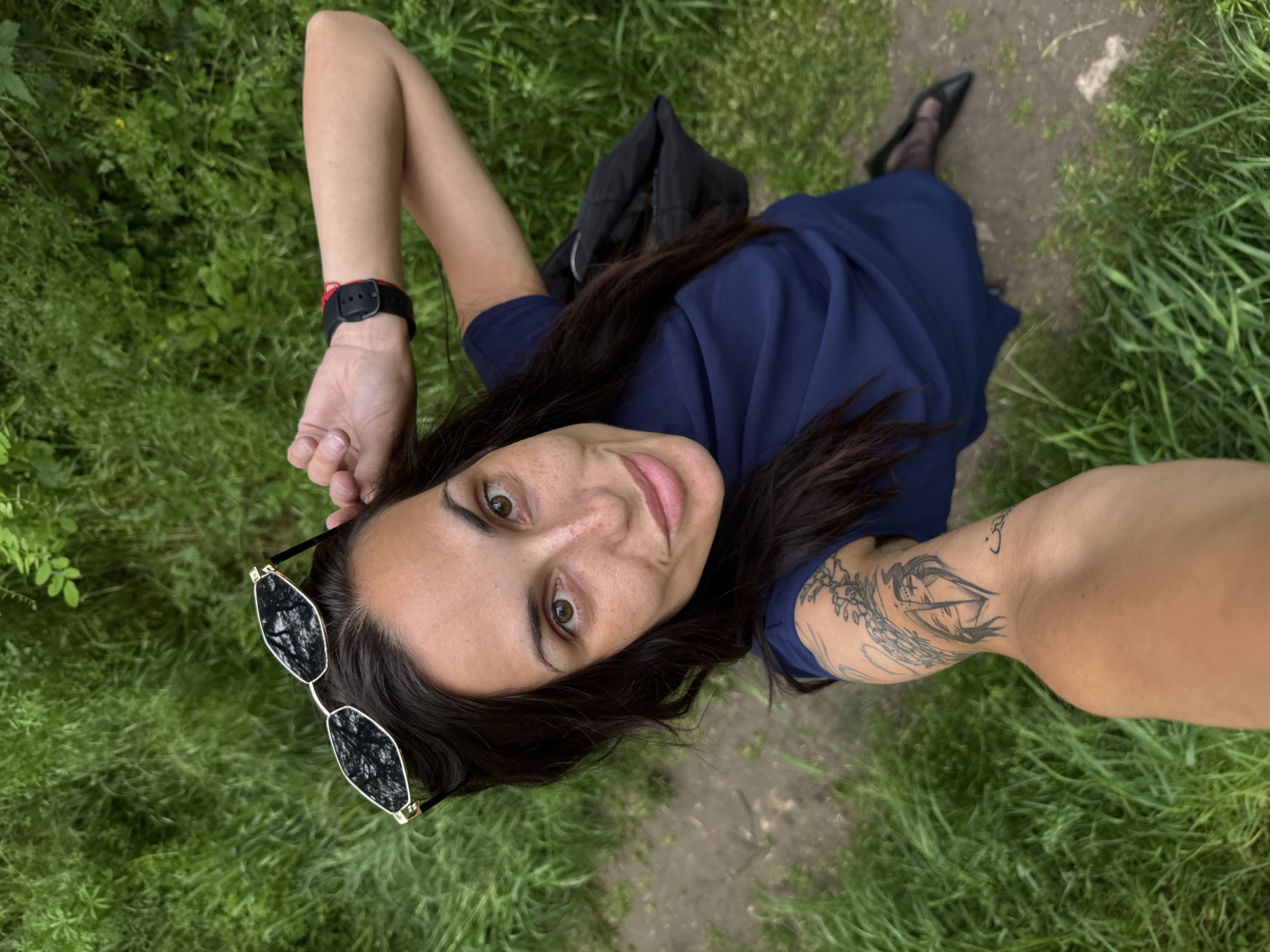 A woman is taking a selfie outdoors. She has dark hair, brown eyes, and fair skin. She's wearing a dark blue dress and black shoes. She has a black watch on her left wrist and a tattoo on her right arm. Sunglasses are resting on the grass near her head. A black bag is visible behind her shoulder. The background consists of green grass and a dirt path. There is no indication of this photo being taken in Pleven, Bulgaria.