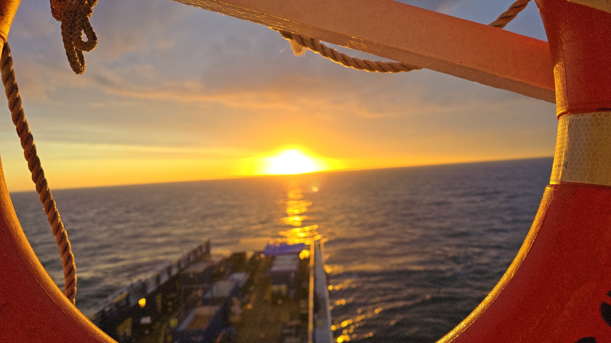 A vibrant sunset is observed through the opening of a life preserver. The sun is positioned on the horizon, casting a bright reflection onto the water. The deck of a ship is visible in the foreground. A rope is attached to the life preserver. The location is Unknown, Unknown.