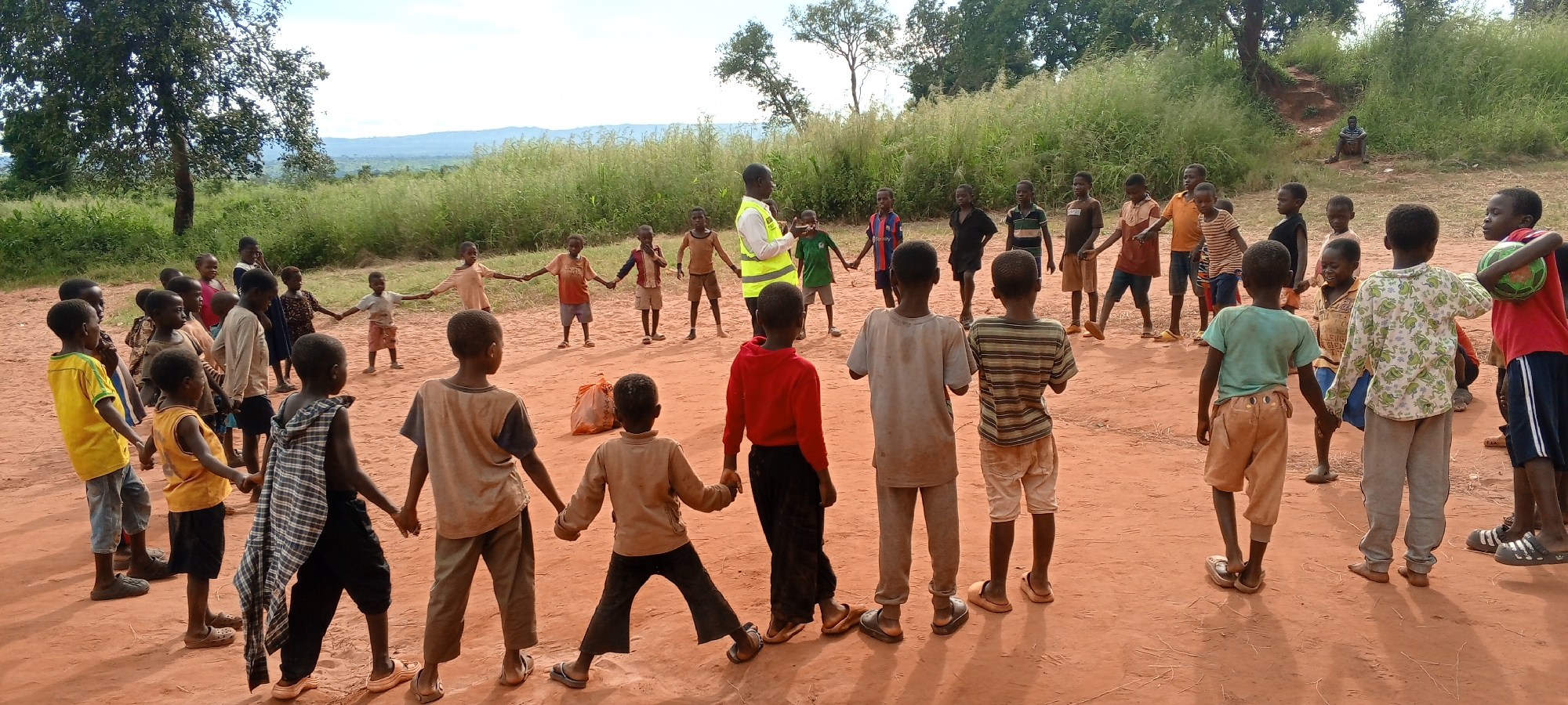 A group of children, seemingly in the city of Ngalula, Democratic Republic of the Congo, are standing in a circle on a red dirt ground. An adult, wearing a yellow reflective vest, stands near one side of the circle. Most of the children are holding hands. The environment consists of trees and vegetation in the background. A small hill and what appears to be a figure sitting on it can be seen in the background.