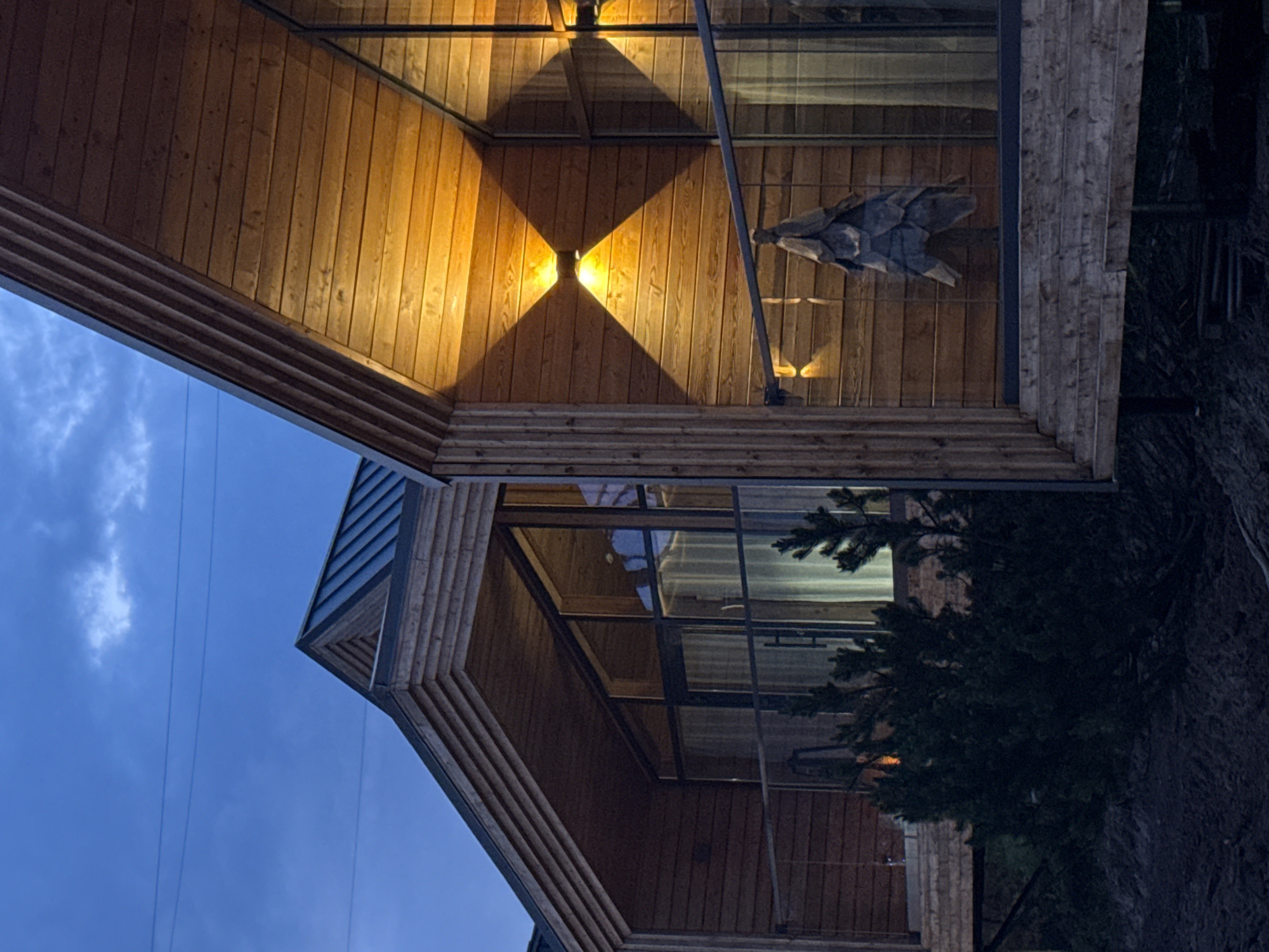 Two modern wooden cabins with large glass windows are situated in Votkinsk, Russia. The sky is dark blue with a few clouds, suggesting it is either dawn or dusk. One cabin, in the foreground, has two exterior lights illuminating the wooden facade and casting diamond-shaped shadows. A decorative wooden object resembling a Christmas tree hangs from the railing of the cabin's balcony. The other cabin, slightly further back and to the left, also features large windows and a similar wooden exterior. A small evergreen tree is visible in front of the second cabin. The ground in front of the cabins appears to be a mix of soil and dark material. Electrical wires are visible against the sky in the background.