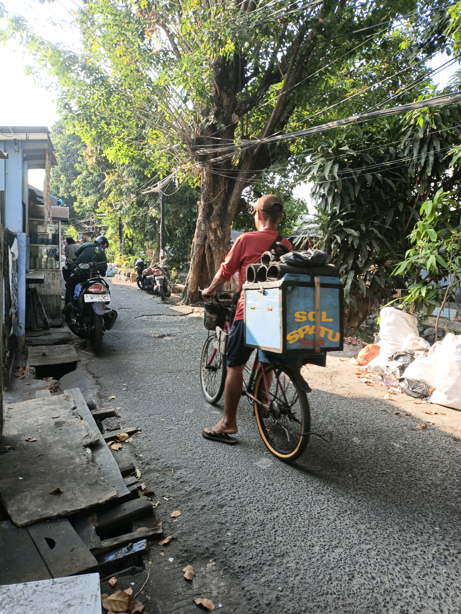 A man wearing a red shirt and a brown baseball cap rides a bicycle down a narrow, asphalt street in West Jakarta, Indonesia. He has a blue box mounted on the back of his bicycle, labeled "SOL SPATU SMR". The box contains several large, cylindrical objects. The street is lined with buildings on the left and trees on the right. Several motorcycles are parked along the left side of the street. There are piles of trash bags on the right side of the street, near a large tree. The sun is shining, casting shadows on the street.