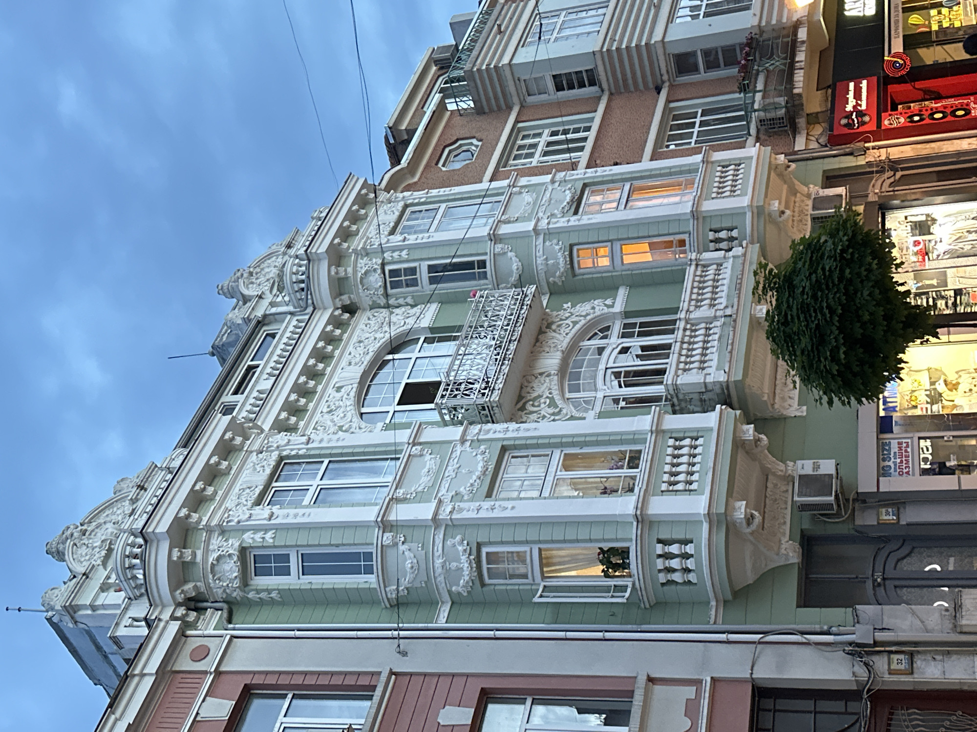 An eye-level, low-angle shot captures the ornate facade of a multi-story building, likely in Varna, Bulgaria, during dusk or early evening. The sky is a cloudy or overcast blue, with several dark utility wires crisscrossing the upper portion of the frame.
The building features intricate architectural details, predominantly in a light green and white/cream color palette. The lower and middle sections are highly decorative, adorned with elaborate stucco carvings, mouldings, and decorative pilasters. Windows vary in shape, including large arched windows with detailed frames and rectangular windows, some featuring small Juliette balconies with metal railings or balustrades. Several windows show warm interior lights on. The upper floors of the building exhibit a more modern, simpler design with flat, rectangular windows and brick-textured walls in reddish-brown tones.
On the right side of the image, the ground level and lower floors show illuminated shopfronts. A dense, dark green tree or bush is visible next to one of these shops. Visible text on a shop sign includes "BIG SIZE" and "БОЛЬШИЕ РАЗМЕРЫ" (meaning "Large Sizes" in Cyrillic). Further up, red neon signs display abstract shapes and stylized letters. A small red plaque or carving on the building's facade reads "1941", possibly indicating the construction year. Near the bottom, a white plate with the number "32" is affixed to a wall. Air conditioning units are attached to the facade at various points. No people are visible.