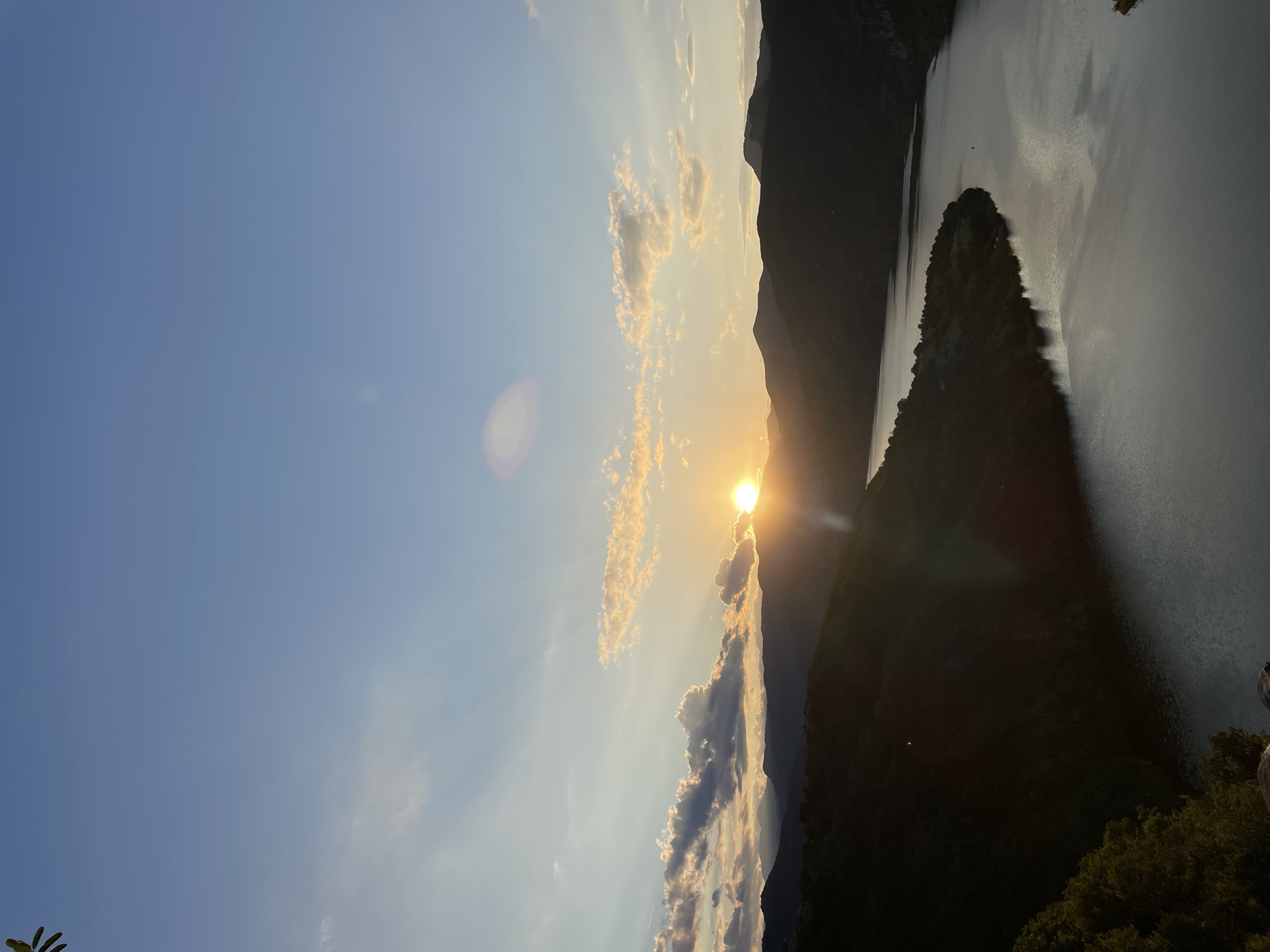 A wide-angle, high-angle shot captures a scenic sunset over a mountainous landscape and a body of water, presumably the Borovitsa Dam, near Borovitsa, Bulgaria. The sun is low on the horizon, partially obscured by clouds and mountains, casting warm golden light across the sky and illuminating the clouds with orange and pink hues. Rays of light emanate from the sun, creating a dramatic effect. Silhouetted mountain ranges form the backdrop, with one prominent peak on the right side of the frame. A large body of water, likely a reservoir or dam, occupies the lower portion of the image, reflecting the twilight sky. The shores of the water are lined with dark, tree-covered hills. The sky is a gradient of blue, transitioning to lighter tones near the horizon. There are no people or discernible man-made structures in the visible area of the image.