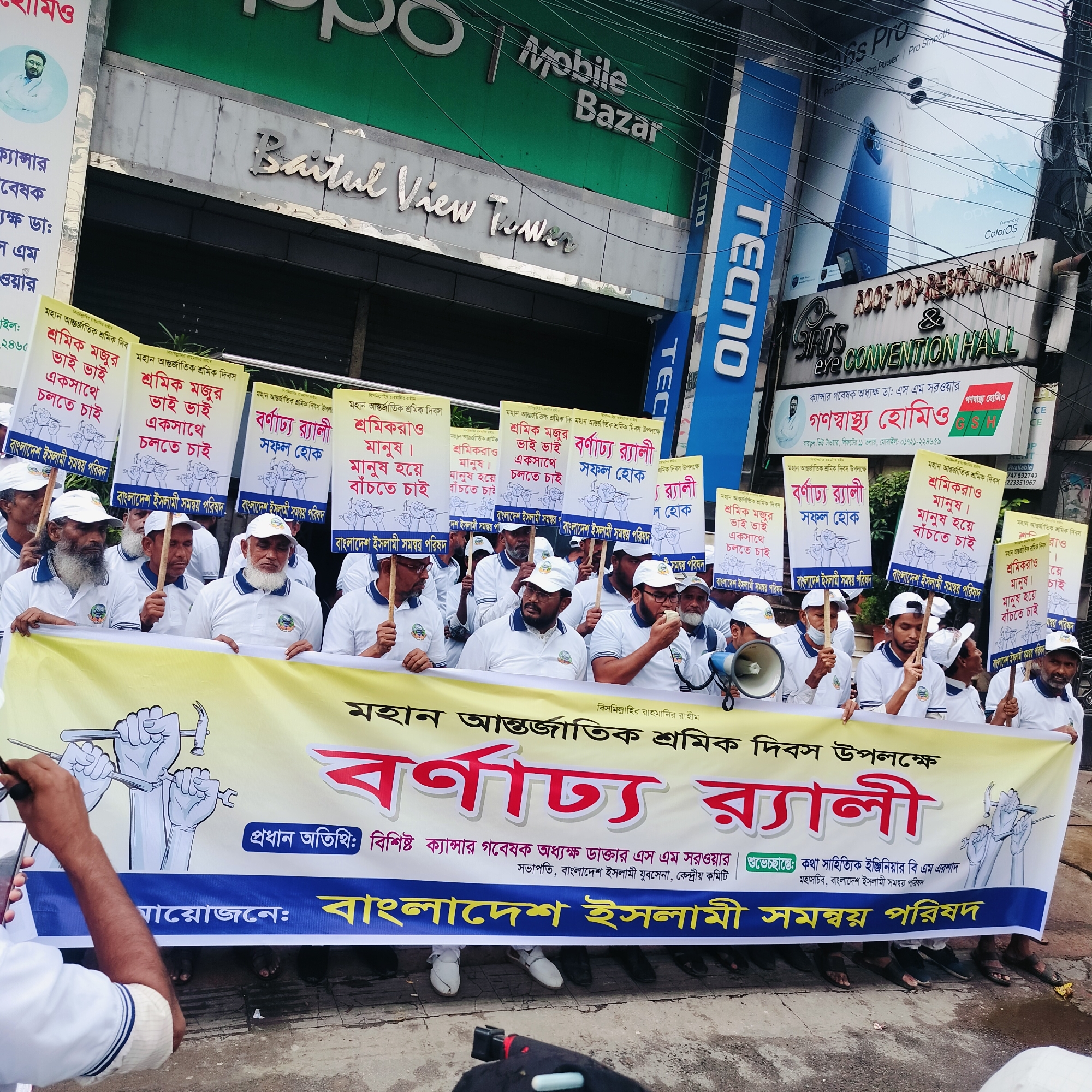 A daytime street demonstration is taking place in Dhaka, Bangladesh, featuring a group of men, most of whom are wearing white shirts and caps, marching with signs and banners. The signs, written in Bengali, mention "International Workers' Day" and advocate for worker unity and rights. A large banner at the front identifies the event as a rally organized by the "Bangladesh Islami Somonnoy Parishad."
The backdrop includes commercial buildings with visible signage, including "Mobile Bazar" and "Baitul View Tower." Other signs display advertisements for "TECNO" mobile phones and “Bird’s Eye Convention Hall”, and Ganoshajjo Homio, a homeopathic medical practice. Power lines and tangled wires are overhead.
