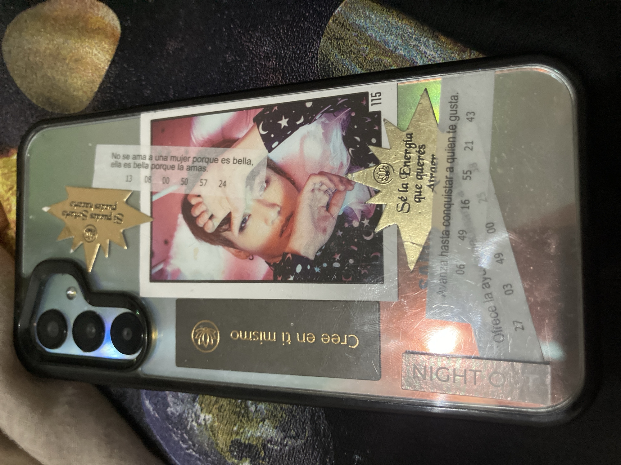 The image displays a smartphone encased in a transparent protective cover, resting on an unidentifiable dark, textured surface. The phone is oriented diagonally, with its camera module visible in the top-left corner, featuring three lenses and a flash.
The clear phone case holds various paper cutouts, stickers, and a photograph, creating a personalized collage effect. A central polaroid-style photo features a young person with light skin and dark hair, looking towards the right side of the frame. They are wearing a dark garment adorned with white stars and crescent moons.
Around the photo and within the case, several pieces of text are visible:
* A white strip on the left reads: "No se ama a una mujer porque es bella, ella es bella porque la amas." (One does not love a woman because she is beautiful, she is beautiful because you love her.) Below this are numbers: "13 08 00 50 57 24".
* A golden star-shaped sticker on the left states: "Si puedes Soñarlo puedes..." (If you can dream it, you can...).
* Another golden star-shaped sticker on the right reads: "Sé la Energía que querés Atraer" (Be the energy you want to attract). A lotus flower logo is also present on this sticker.
* A white strip near the top right includes text: "Avanza hasta conquistar a quien te gusta." (Advance until you conquer the one you like.) and numbers: "06 49 16 55 21 43".
* Further down on the right, a partial strip reads: "Ofrece la ay..." (Offer help...) with numbers: "27 03 49 00".
* A dark rectangular piece at the bottom center has golden text: "Cree en ti mismo" (Believe in yourself) with a lotus flower logo.
* At the bottom right, a metallic strip features the embossed text: "NIGHT OUT".
* A small white tab at the top right has the number "115".
The overall scene conveys a sense of personal expression and motivation through quotes and imagery. The lighting is artificial, suggesting an indoor setting.