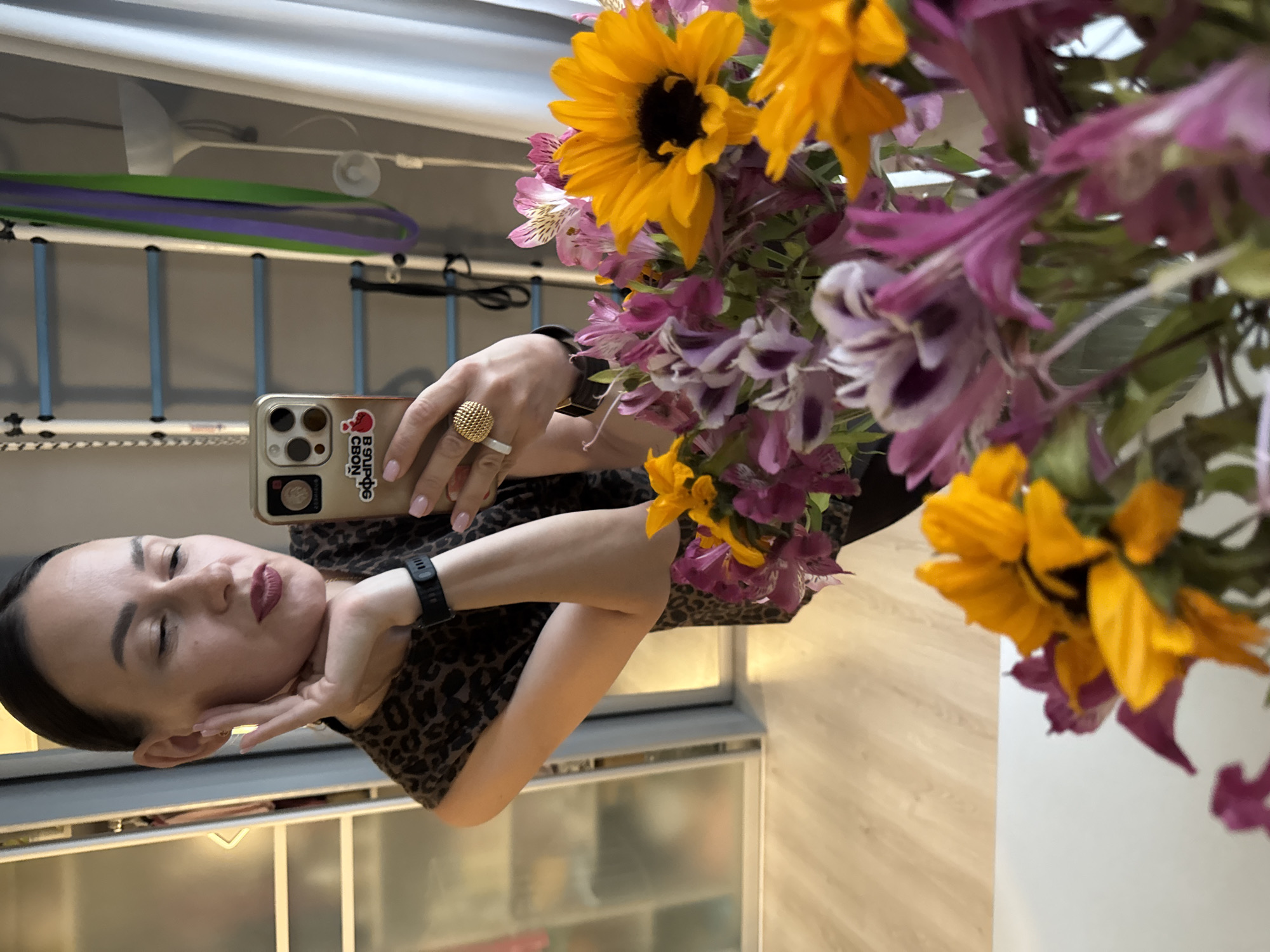 In a well-lit interior, a woman with dark hair pulled back, wearing a leopard-print top and a black smartwatch, is seen taking a selfie in a mirror. Her left hand is raised to her cheek, and she is wearing a gold ring with a textured, spherical design on her right index finger. In the foreground, a colorful bouquet of flowers, including sunflowers and purple and pink alstroemeria, is visible. In the background, a set of blue and light blue gymnastic bars is mounted on a wall, with purple and green resistance bands hanging from them. A white floor lamp and a white curtain are also present in the room. The floor appears to be light-colored wood laminate. The woman's mobile phone, with a gold case featuring a sticker that reads "ИСПРАВЬ СВОЙ СЛОГ" (Fix your syllable) and a red heart icon, is held in her right hand, capturing the reflection. The overall scene suggests a personal setting, possibly a home or a gym, in Sochi, Russia.