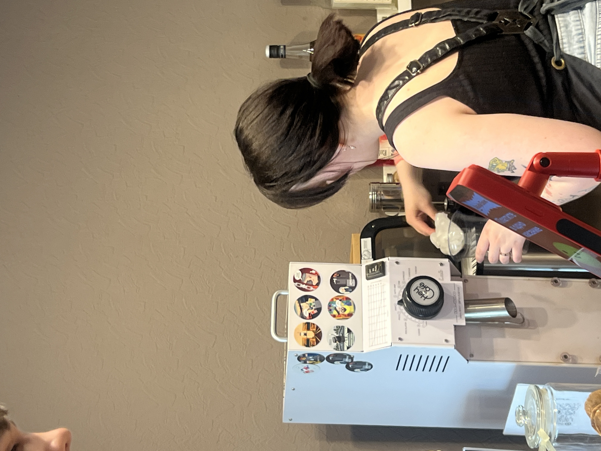 A woman with dark hair in a high ponytail is leaning over a machine with a red handle and a dial labeled "Hey Cake." The machine has a series of circular images on its side, and there is a glass jar with cookies visible in the lower right corner. The background is a plain wall. The scene appears to be indoors, possibly in a bakery or a confectionary. It is unclear if this image was taken in the city of Chaikovsky, Russia.